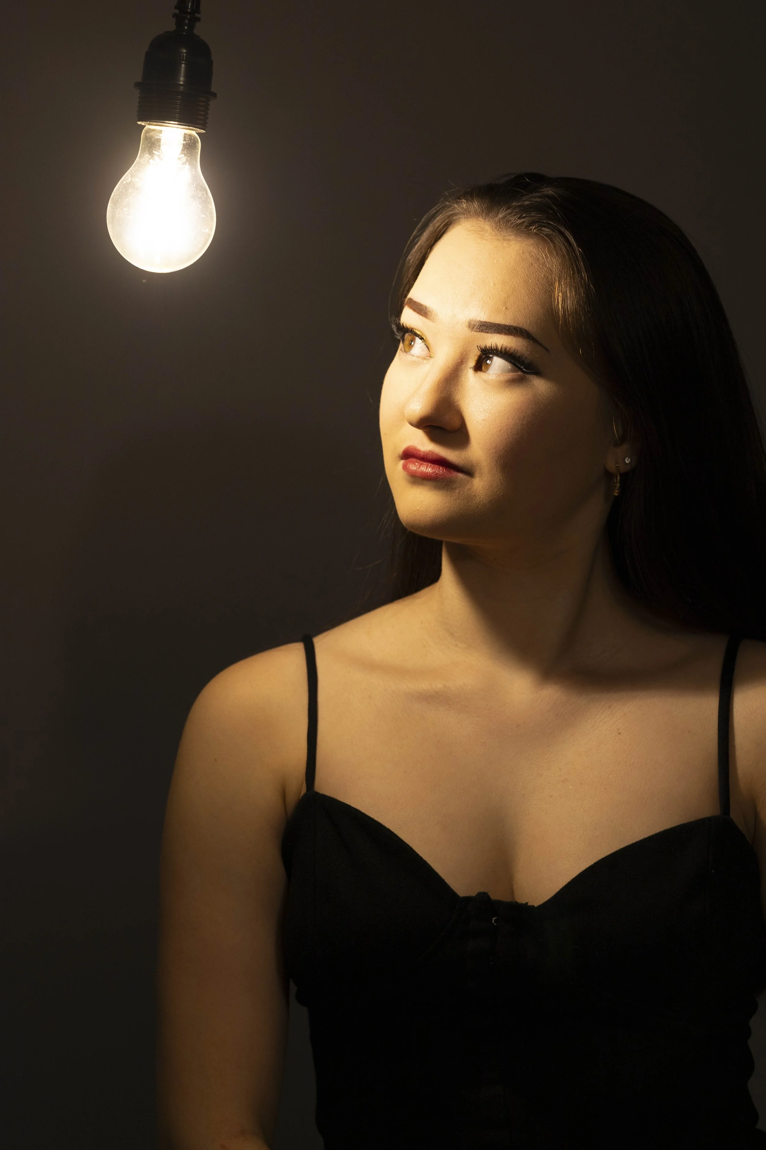 A young woman with dark hair and light skin gazes at a bright light bulb hanging from the ceiling against a dark background.