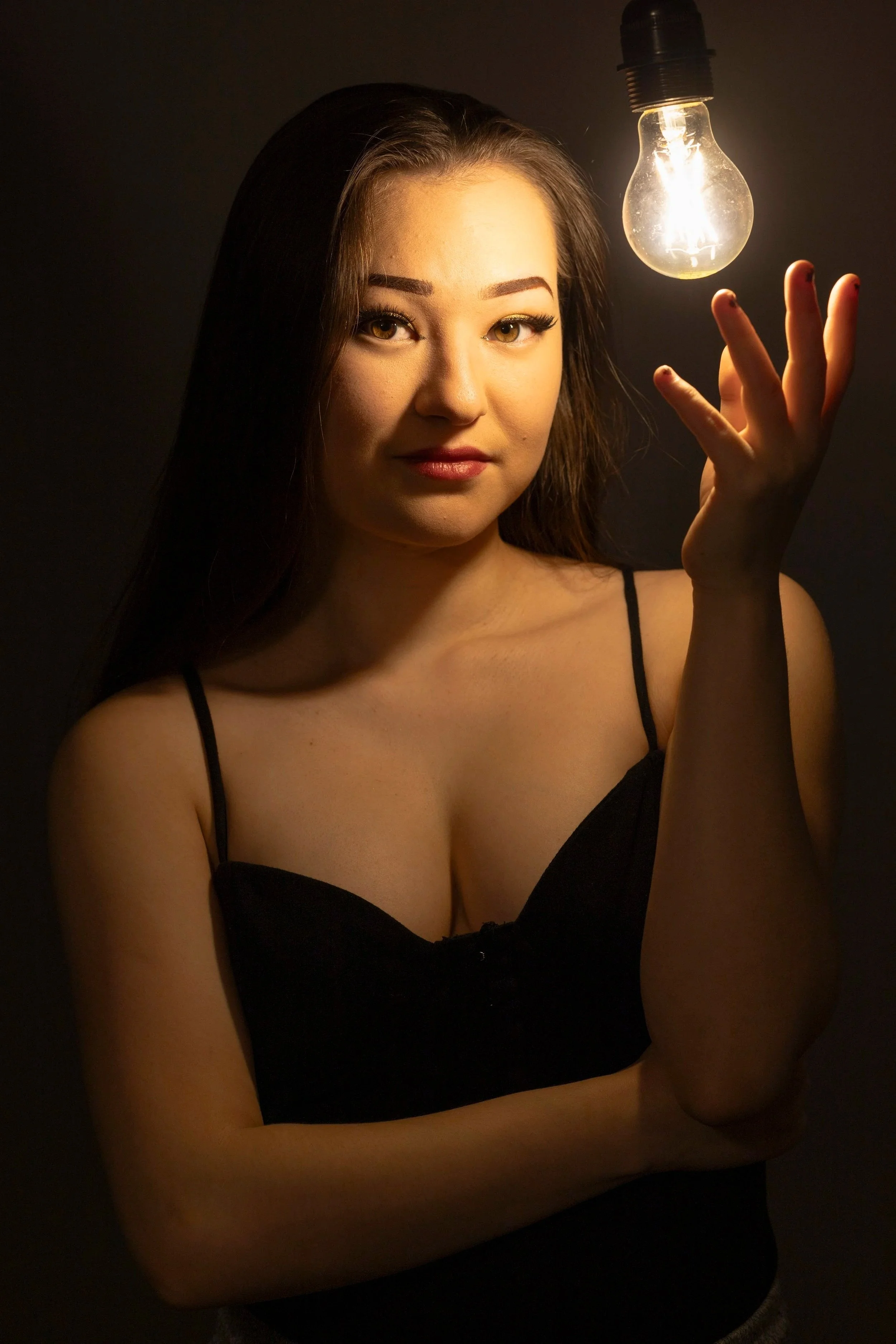 A woman with long dark hair and makeup holds her arm under her chest and reaches out with her right hand to a glowing lightbulb hanging above her head in a dark setting.