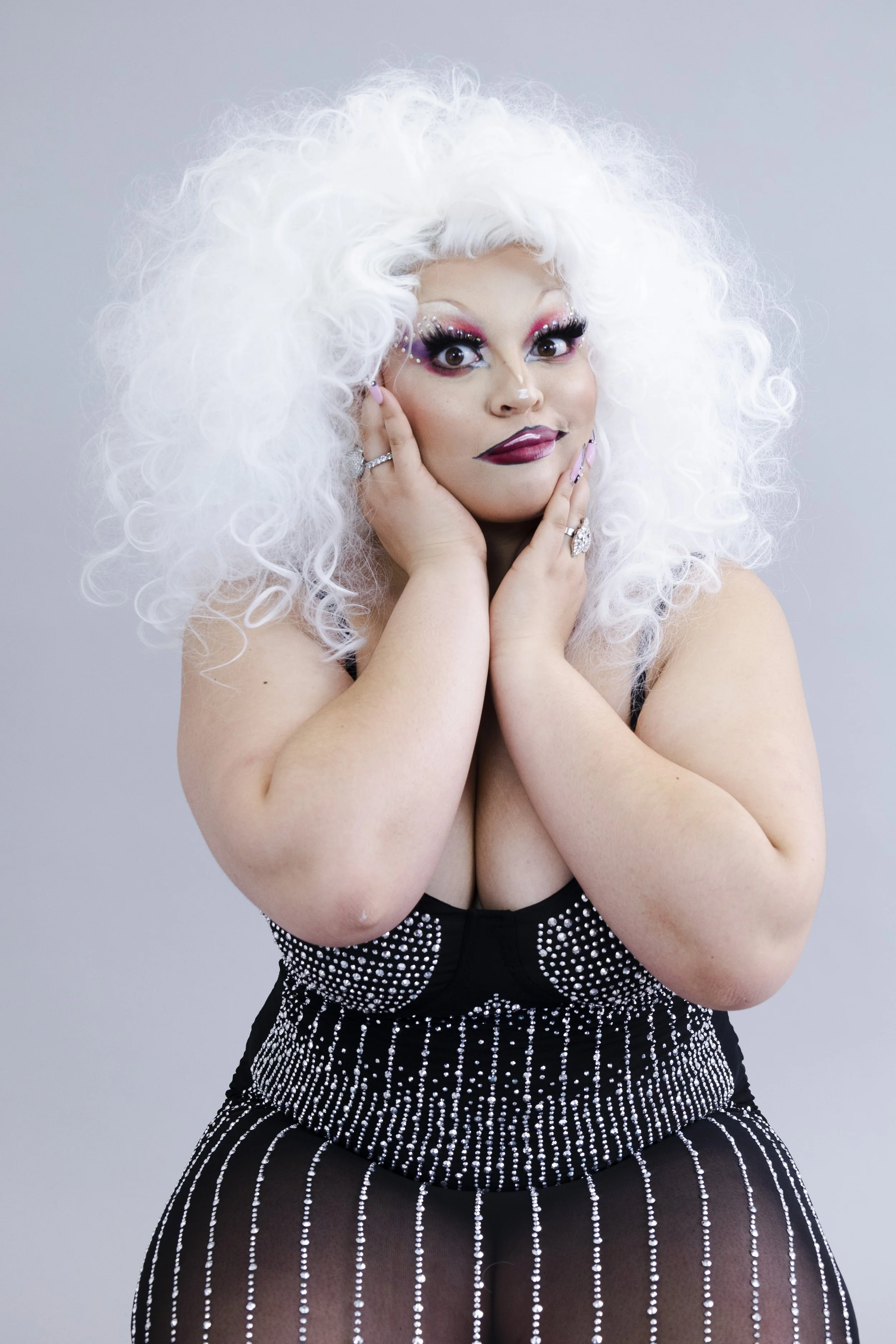 Woman with curly white hair, bold makeup, and jewelry, posing in a black, bejeweled outfit.