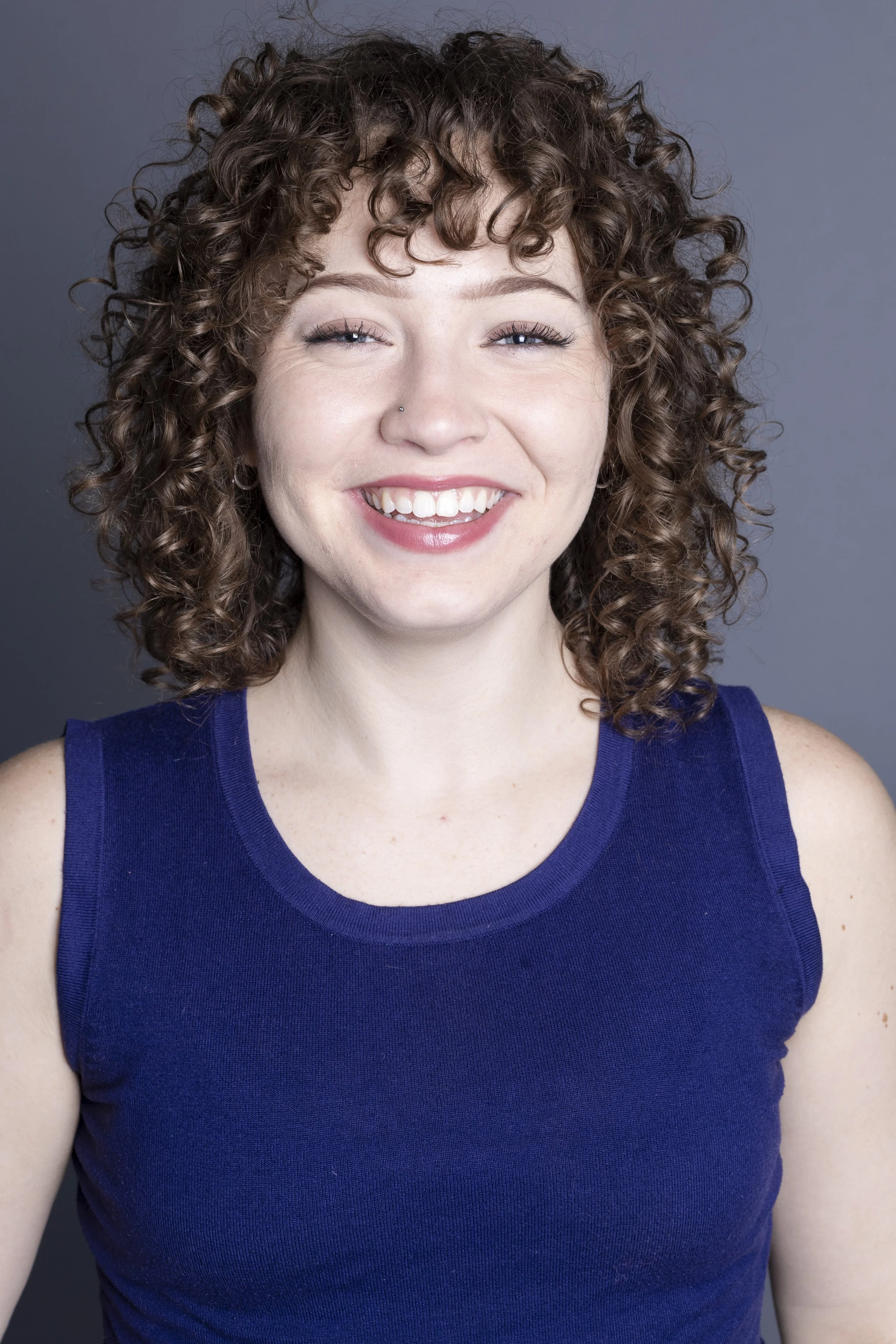 A woman with curly brown hair smiling, wearing a blue sleeveless top, against a gray background.