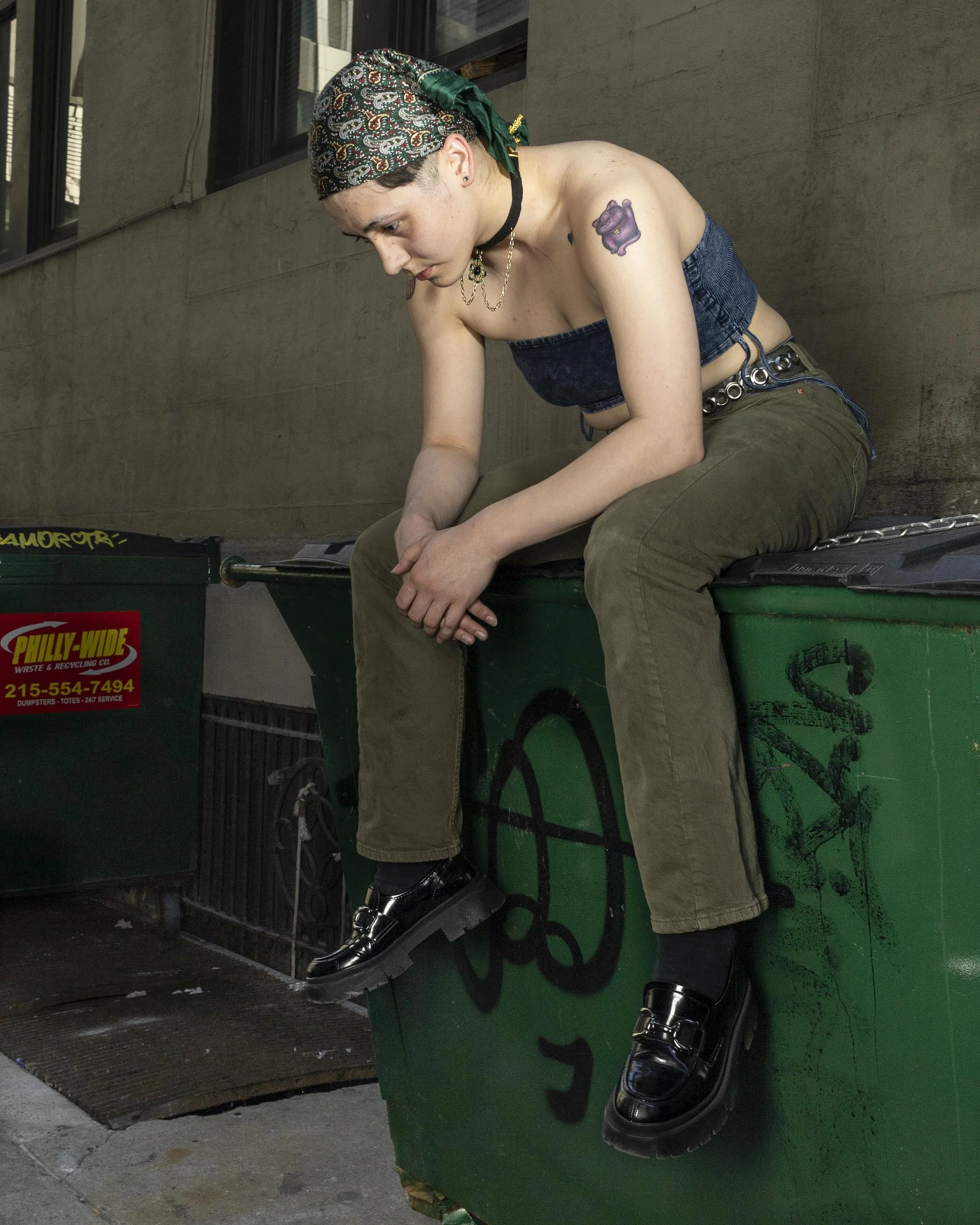 Young woman with a tattoo on her left shoulder, wearing a patterned bandana, a strapless denim top, olive pants, and shiny black loafers, sits on a green dumpster with graffiti, looking downward with her hands clasped.