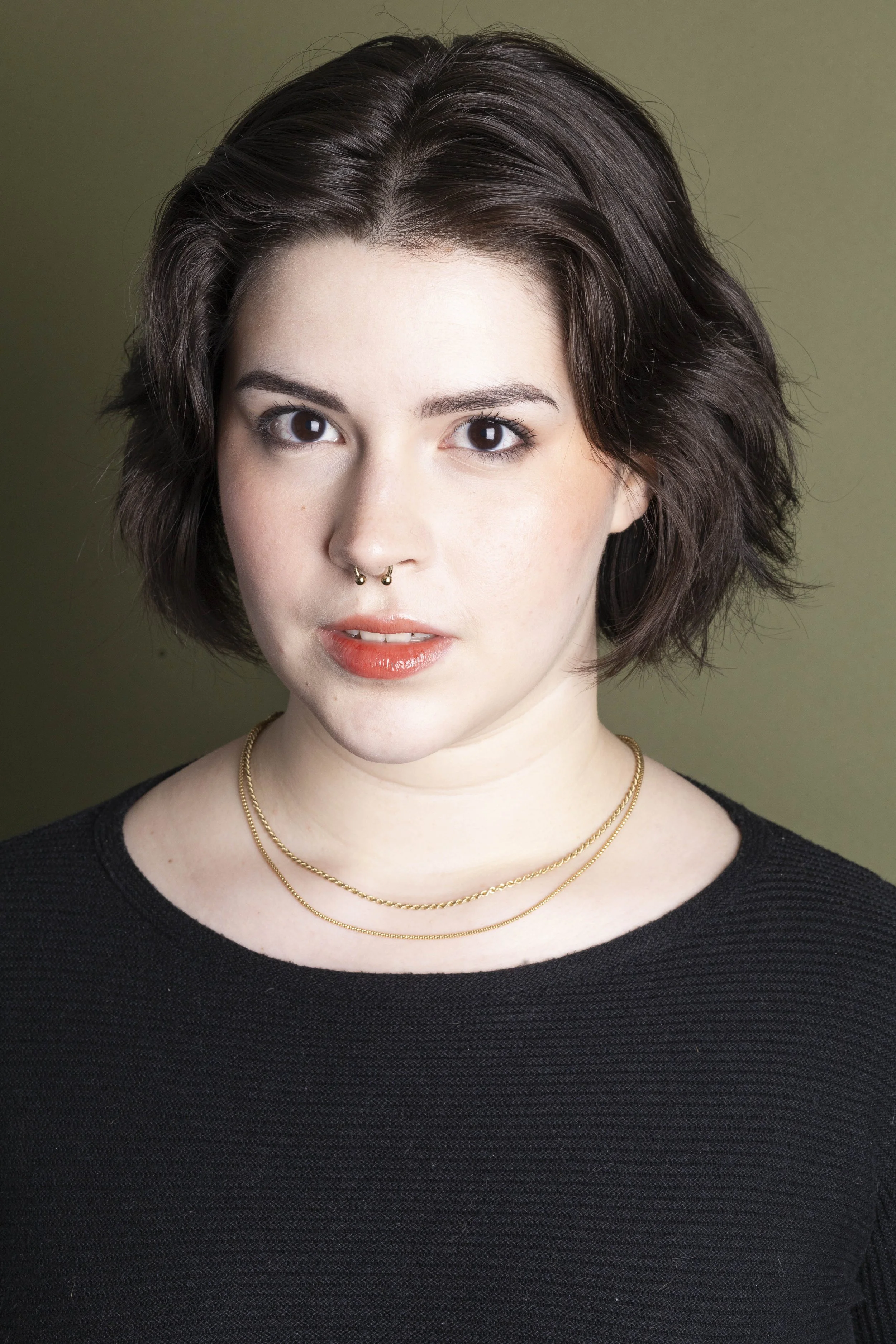 Portrait of a young woman with short wavy dark hair, light skin, and brown eyes, wearing a black top and layered gold necklaces, standing against a plain muted green background.