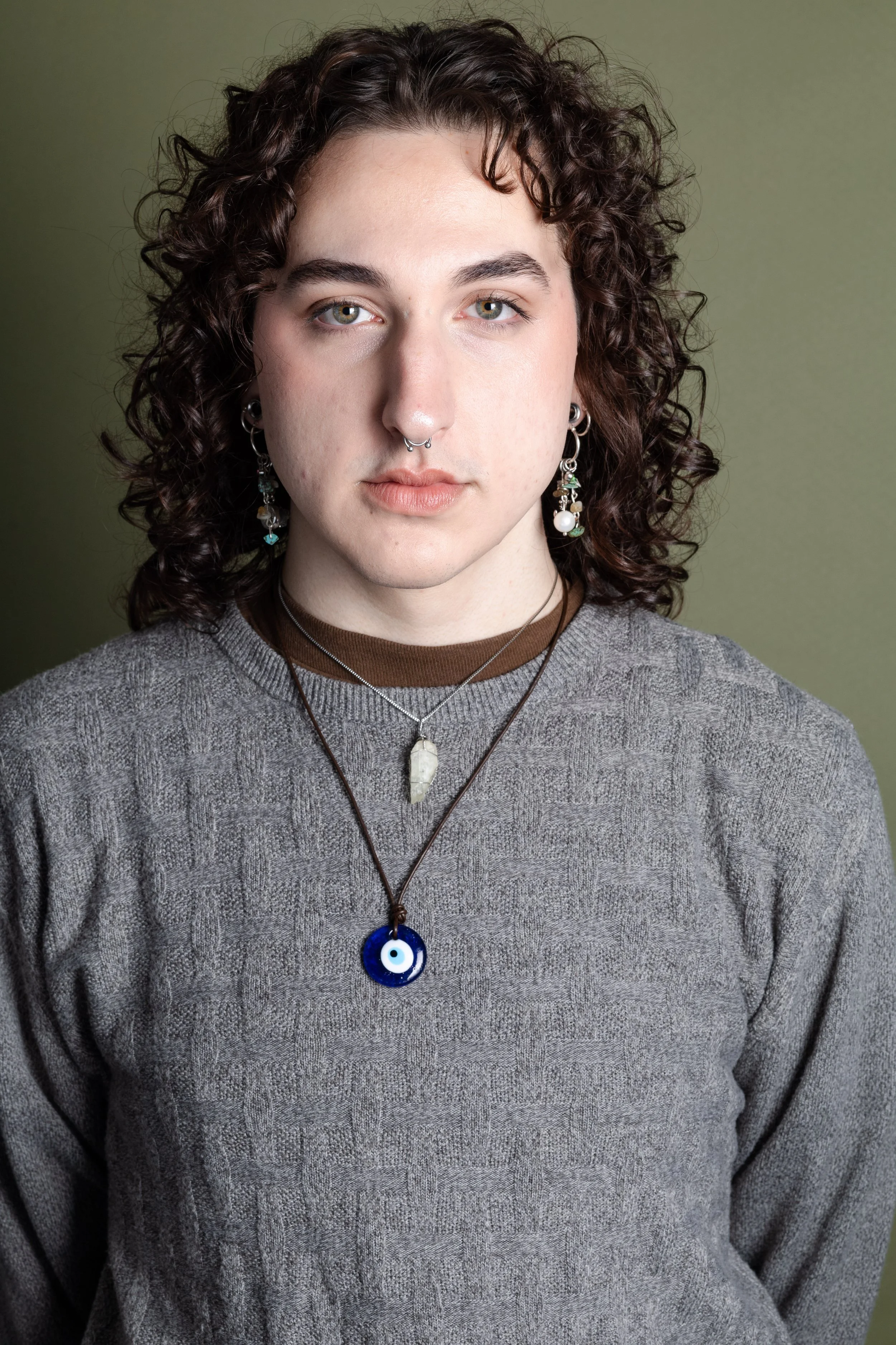 A young person with shoulder-length curly brown hair, light skin, and green eyes, wearing a gray sweater over a brown shirt, with multiple necklaces and earrings, looking directly at the camera against a green background.