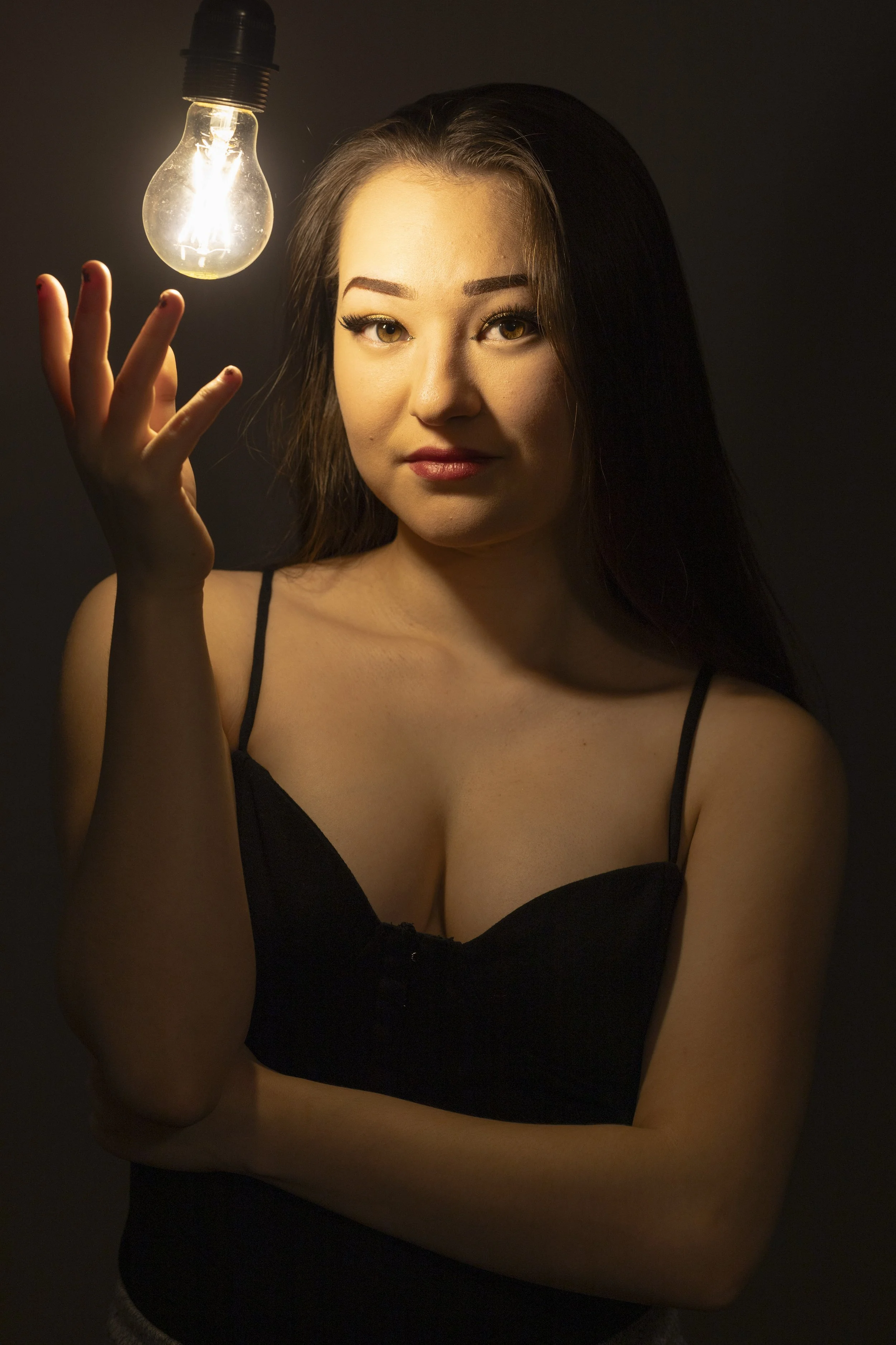 A young woman with long dark hair and light skin holds her hand near a lit lightbulb hanging from the ceiling, wearing a black spaghetti strap top and looking at the camera against a dark background.