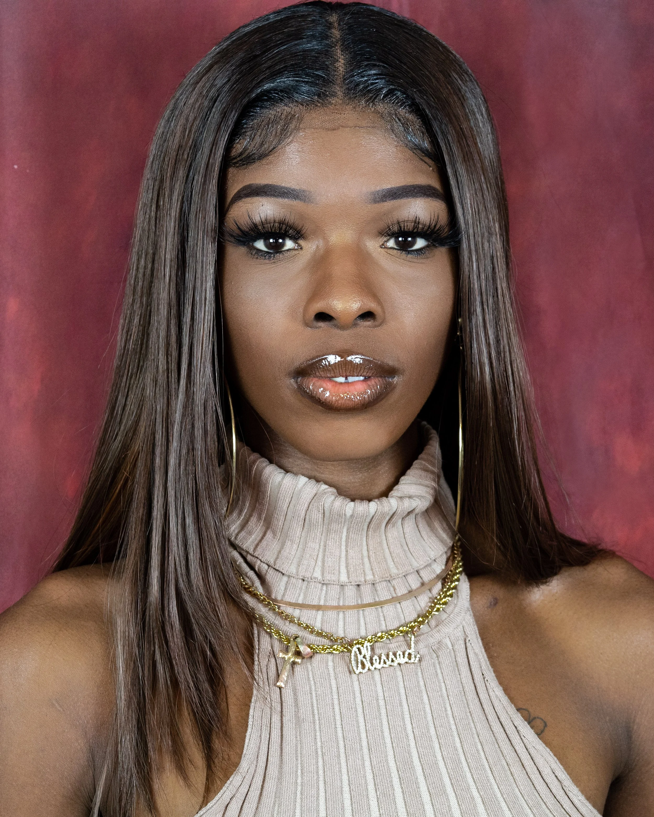 A close-up portrait of a woman with long, straight dark hair, wearing a beige turtleneck top, gold jewelry, and makeup, against a red background.