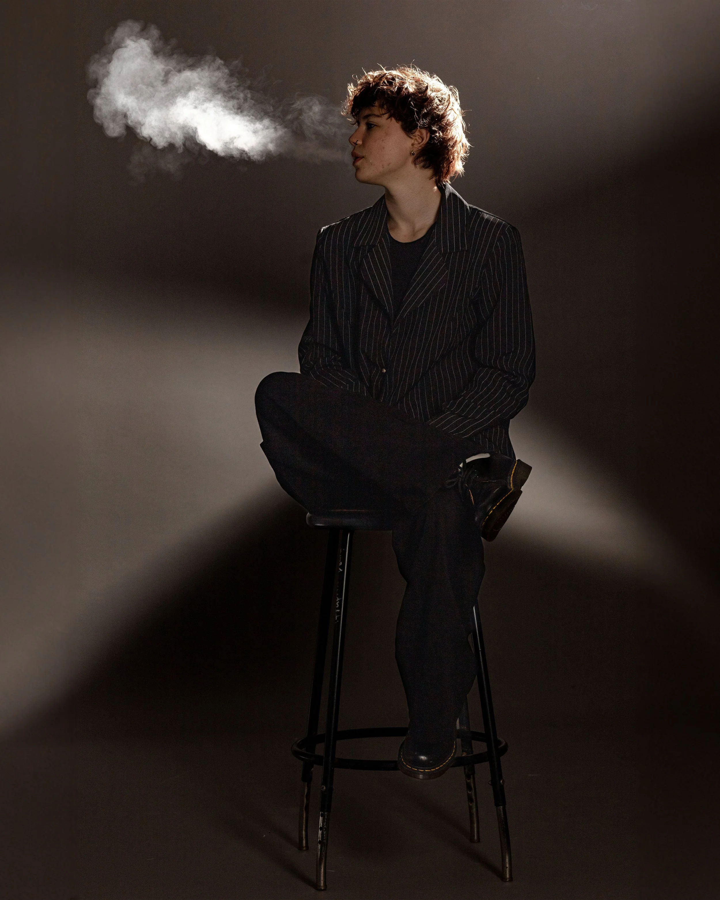 A young person with short, curly hair sitting with one leg crossed over the other on a stool, exhaling vapor or smoke against a dark background.