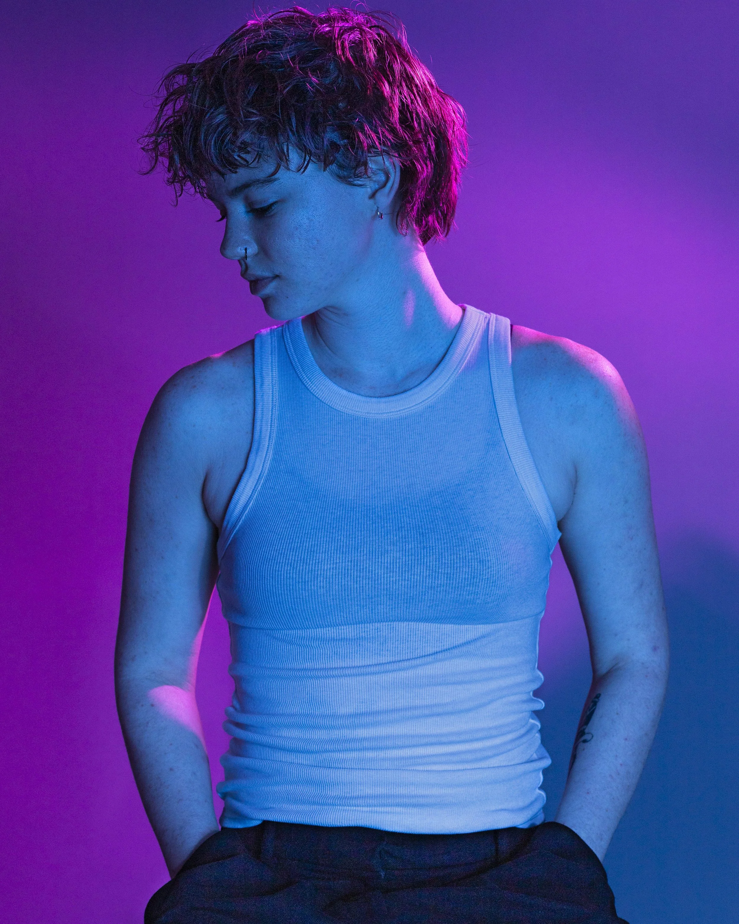 A person with short curly hair and multiple earrings wearing a white tank top, standing against a pink and purple lighting background, looking down with hands in pockets.