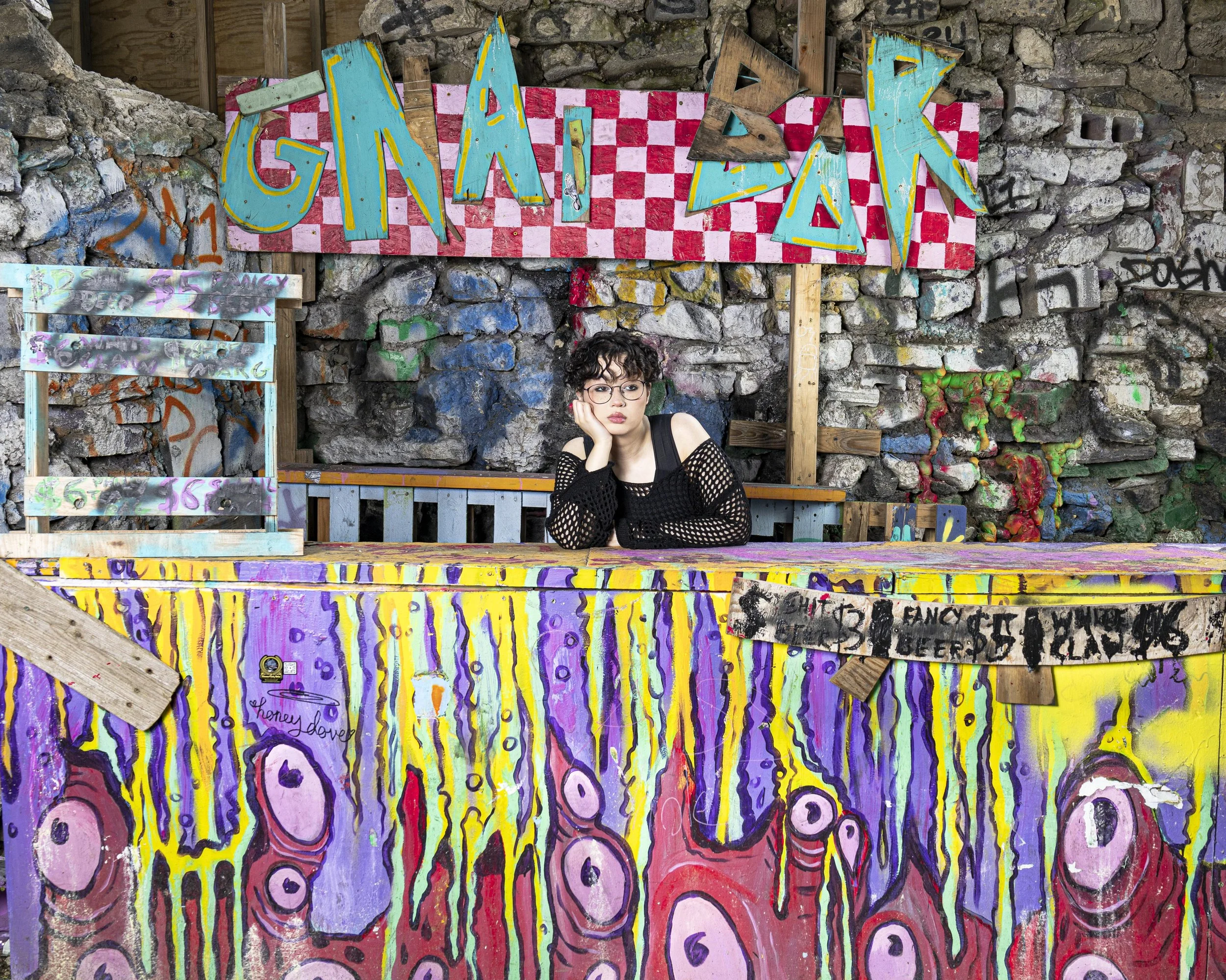 A young woman with short curly hair and glasses sits at a colorful, painted table with her head resting on her hand, in front of a graffiti-covered stone wall with a large, bright, checkered red and pink sign that says 'GNAI DZK' in teal, yellow, and brown letters.