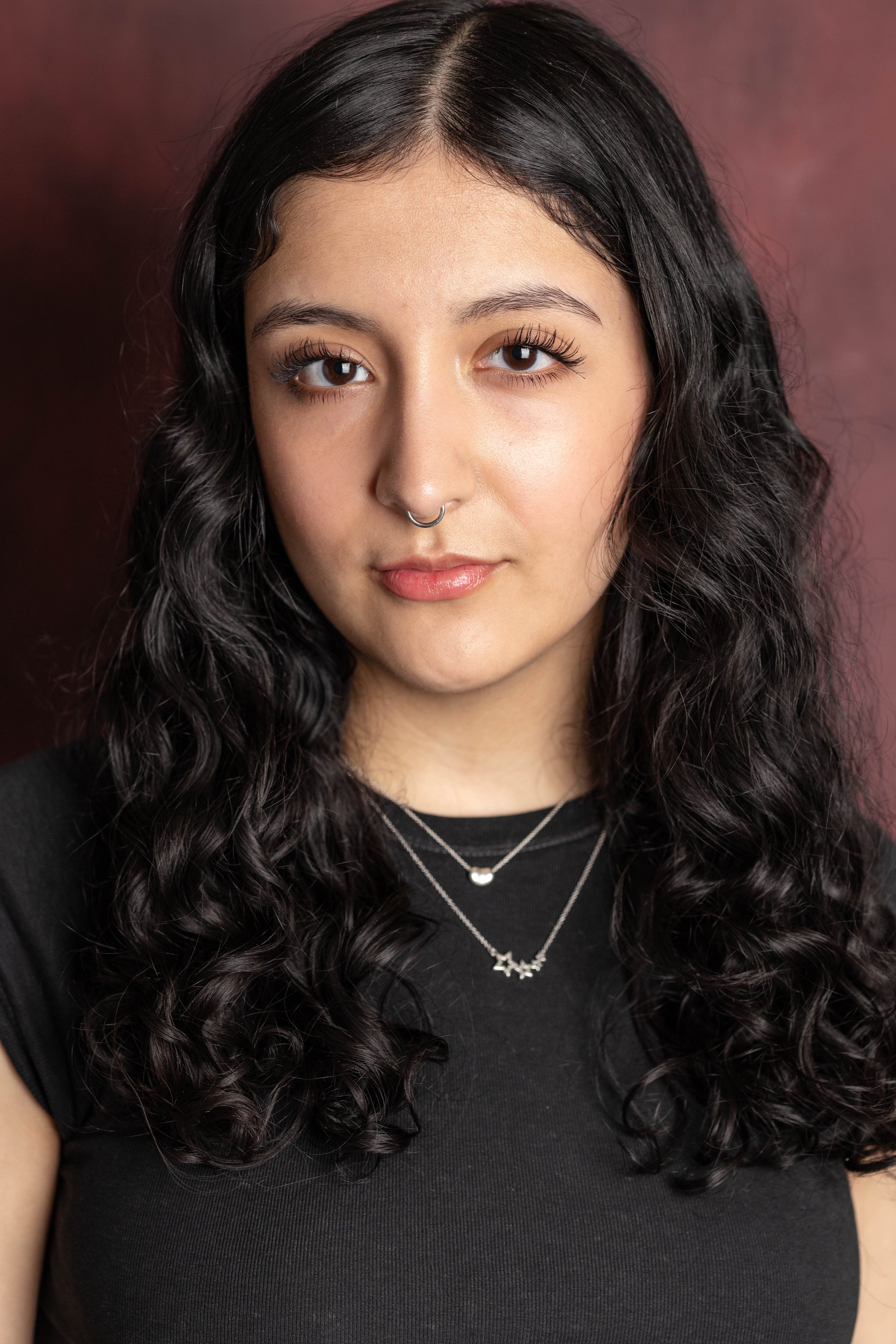 Portrait of a young woman with long curly dark hair, light makeup, a septum piercing, and wearing layered necklaces and a black top, standing against a maroon background.