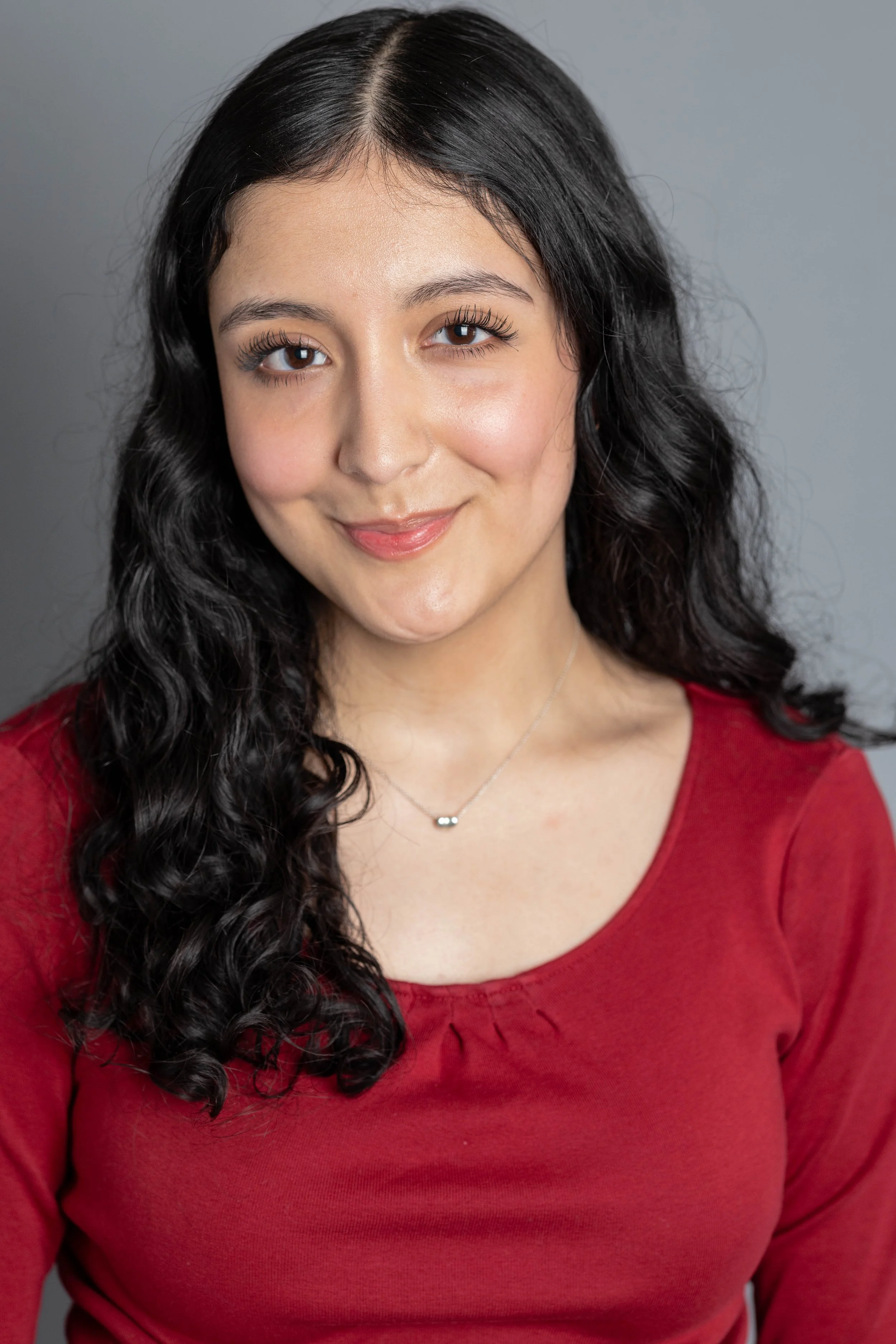 A young woman with long, dark, curly hair wearing a red top and a delicate necklace, smiling at the camera against a gray background.