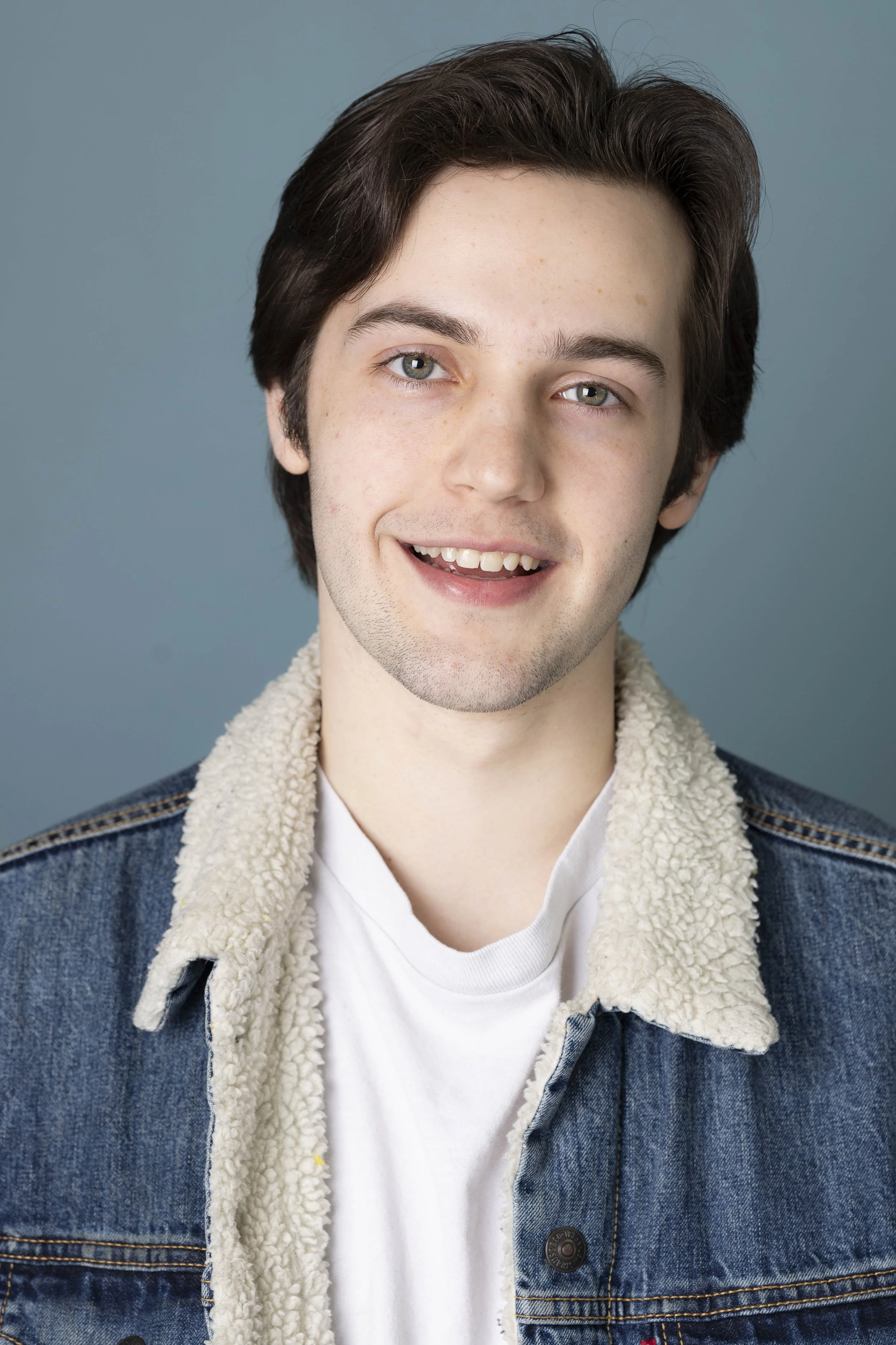 Portrait of a young man with dark hair, blue eyes, smiling, wearing a denim jacket with beige fleece collar, and a white t-shirt against a blue background.