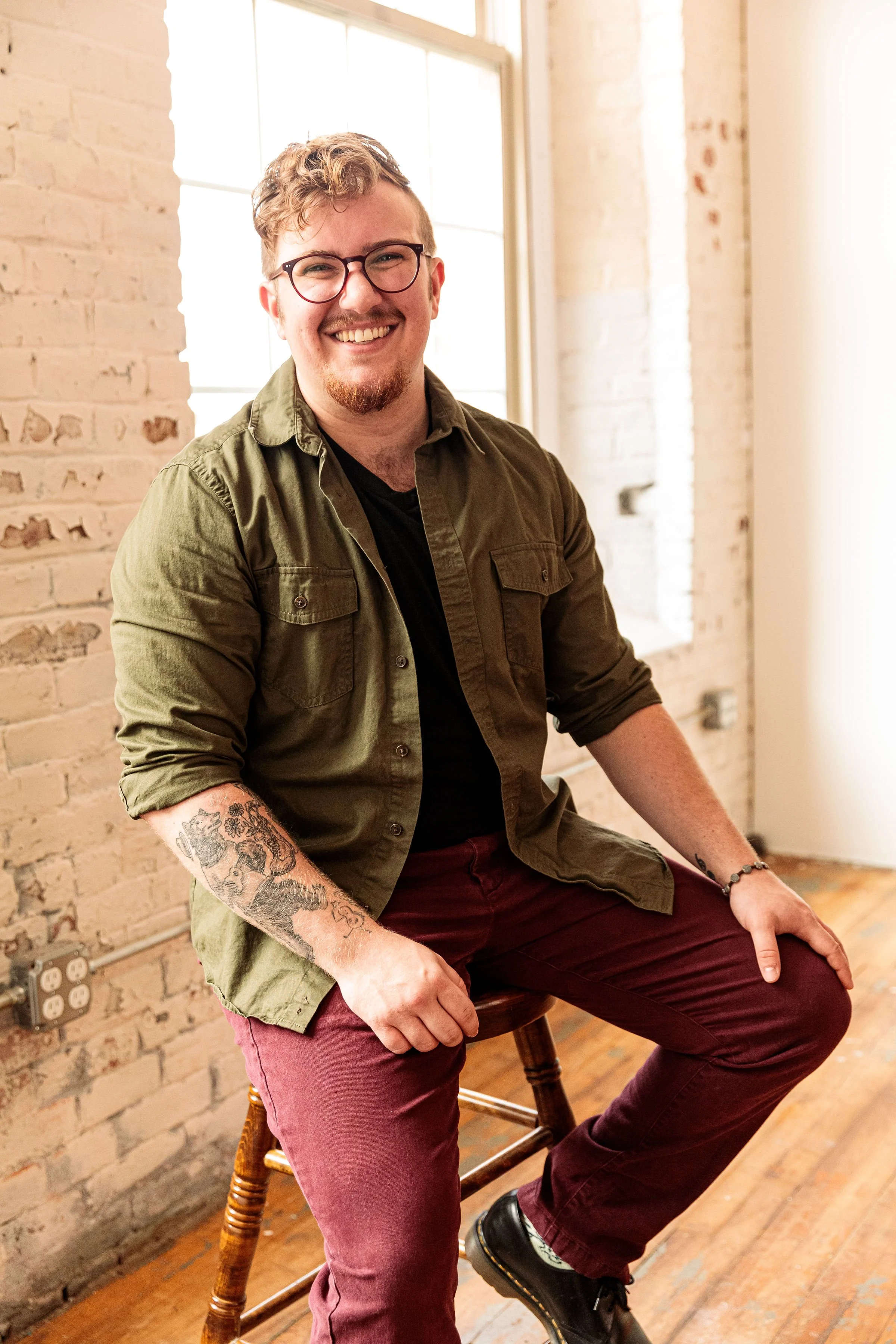 A smiling young man with glasses and tattoos sitting on a wooden stool by a window in a bright room with brick walls.