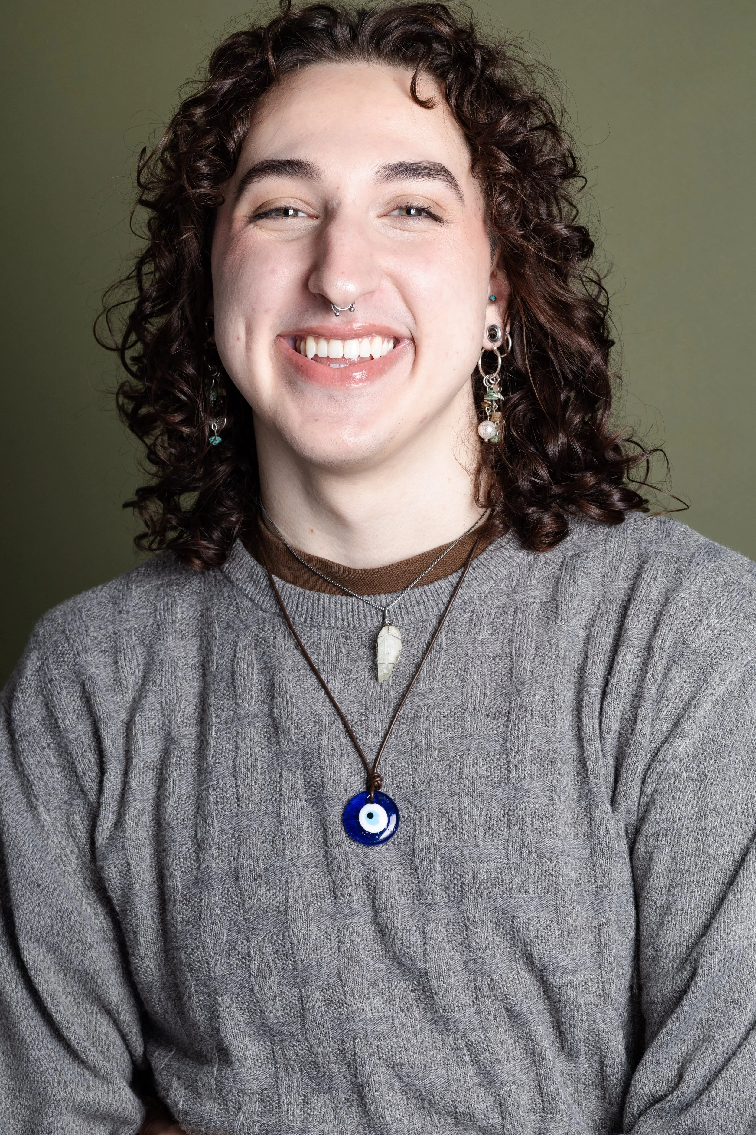 A young person with curly dark hair and light skin, smiling while wearing a gray sweater and multiple necklaces with different pendants, including a blue evil eye pendant, posing against a green background.