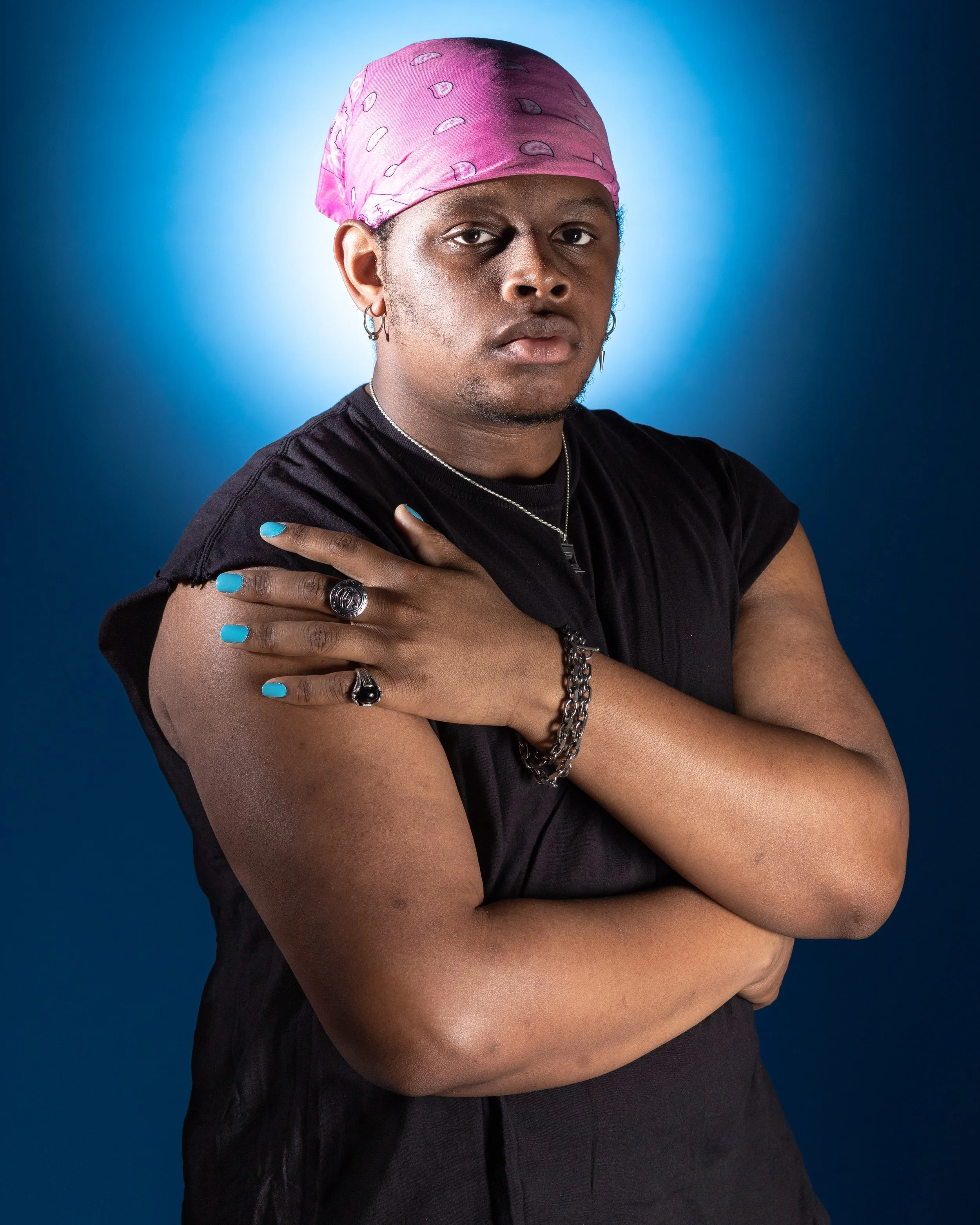 Portrait of a person wearing a pink bandana, black t-shirt, with jewelry, against a blue background.