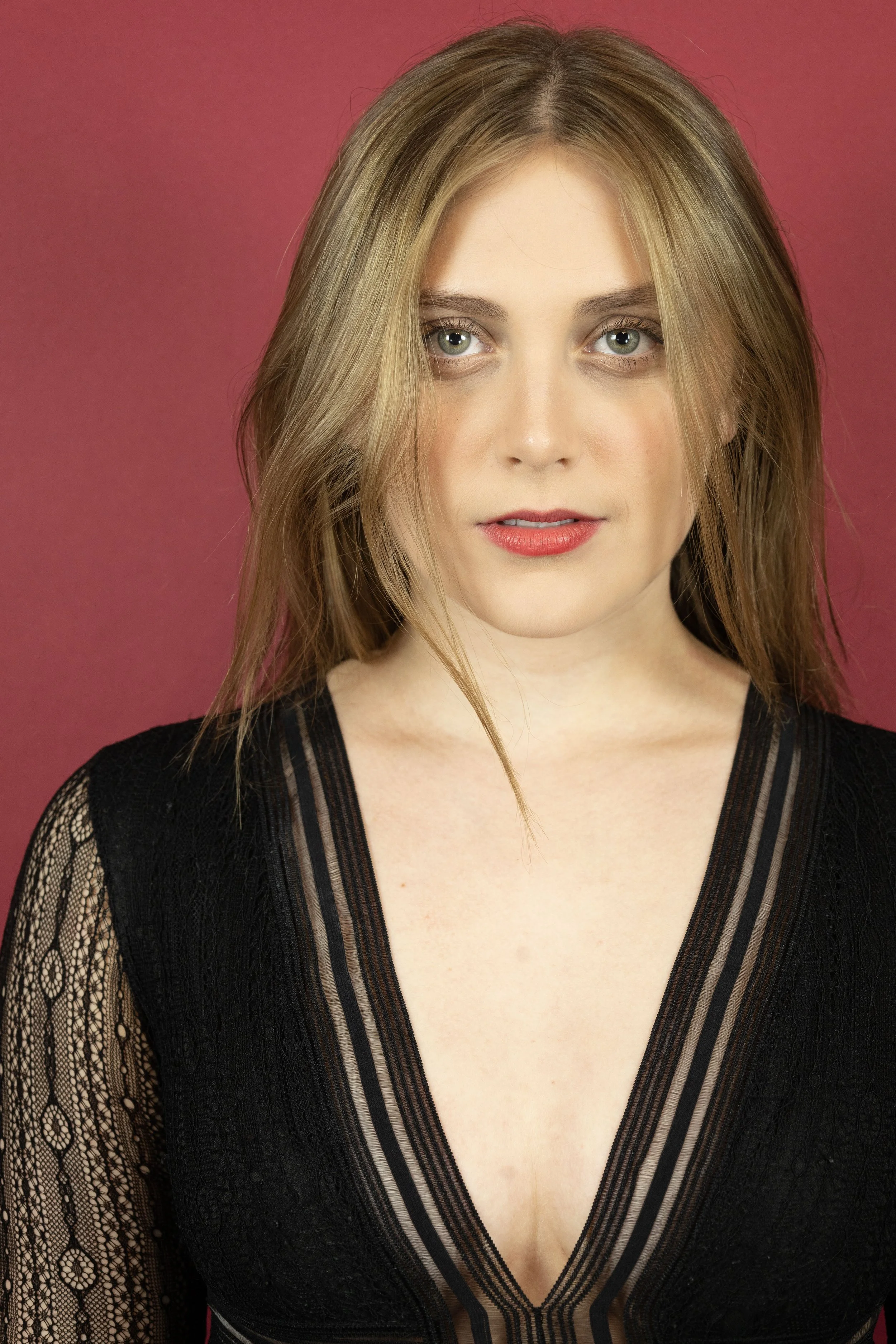 A young woman with blonde hair and green eyes wearing a black lace dress against a pink background.