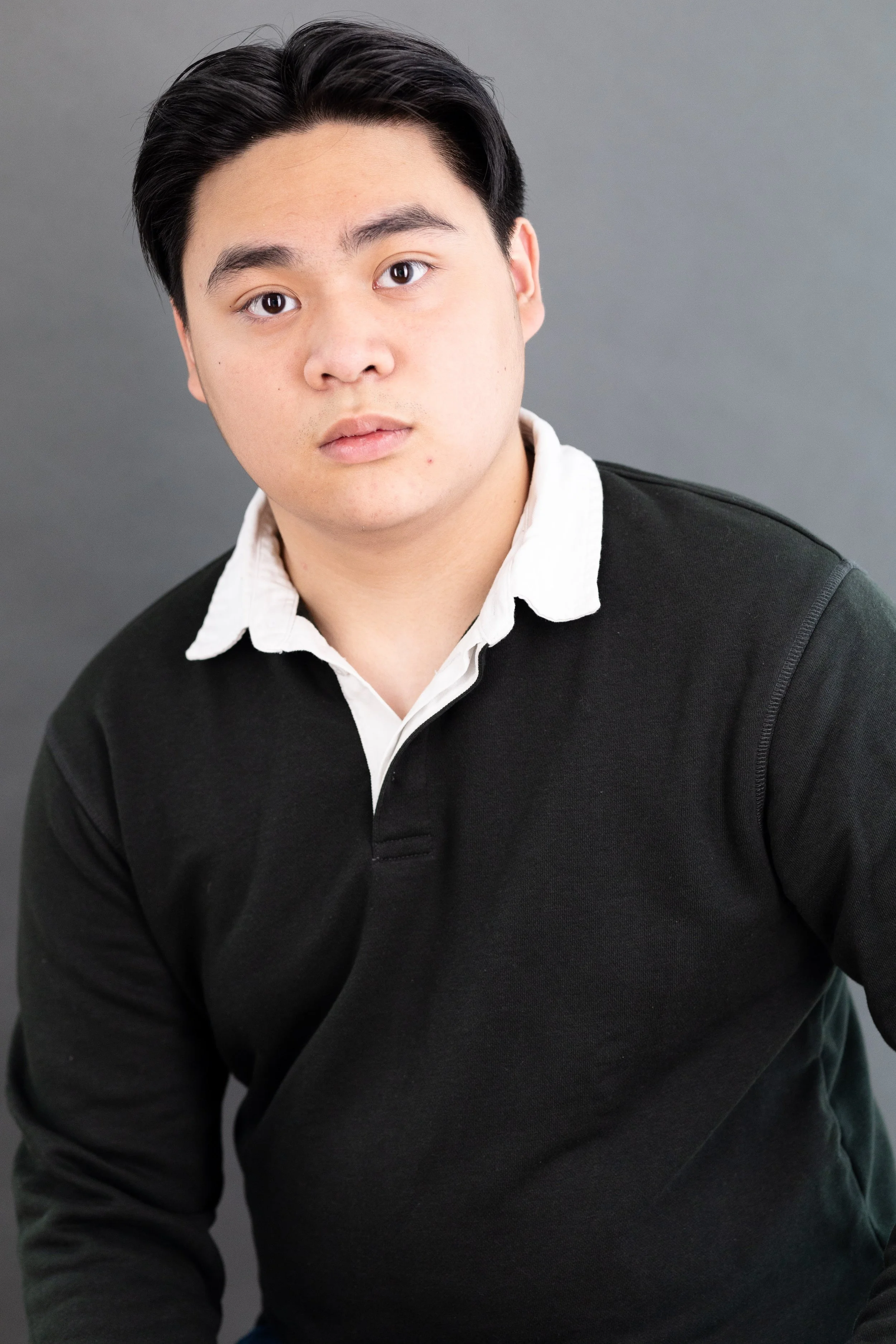 Portrait of a young man with black hair wearing a black sweater over a white collared shirt against a gray background.