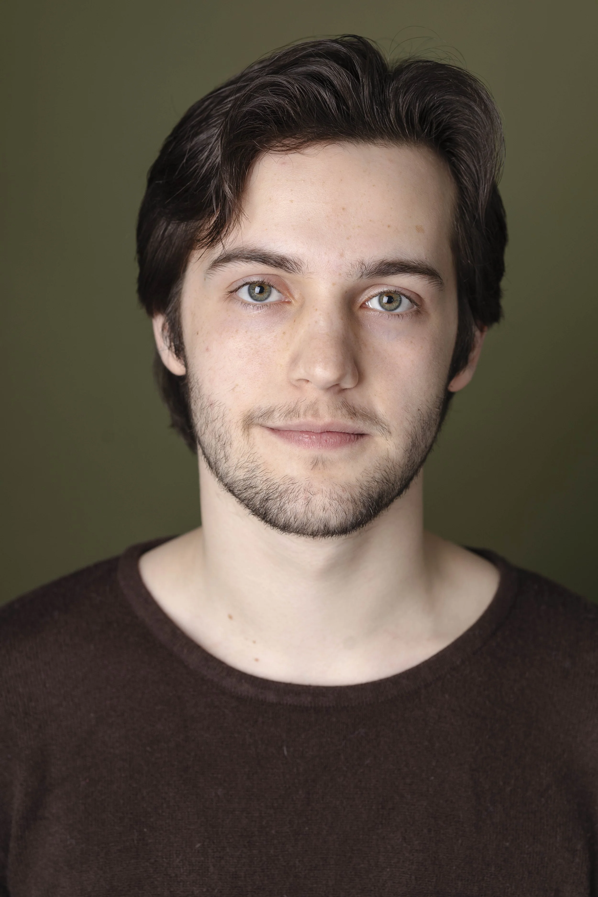 Headshot of a young man with light skin, brown hair, and blue eyes, smiling softly, wearing a dark brown sweater, with a green background.