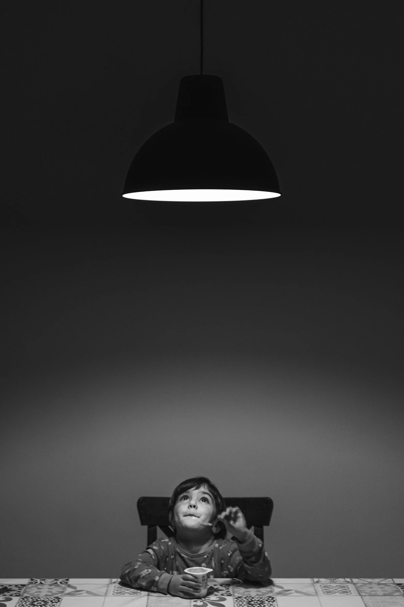 A young child sitting at a dining table looking up at a hanging ceiling lamp with a neutral expression, holding a spoon and a small cup, black and white photo.