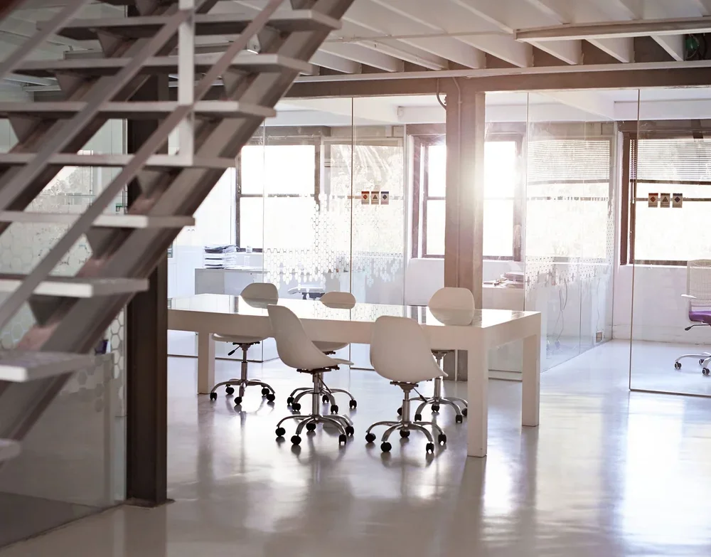 Modern office space with a white table, several white chairs on casters, glass partitions, and large windows with sunlight streaming through.