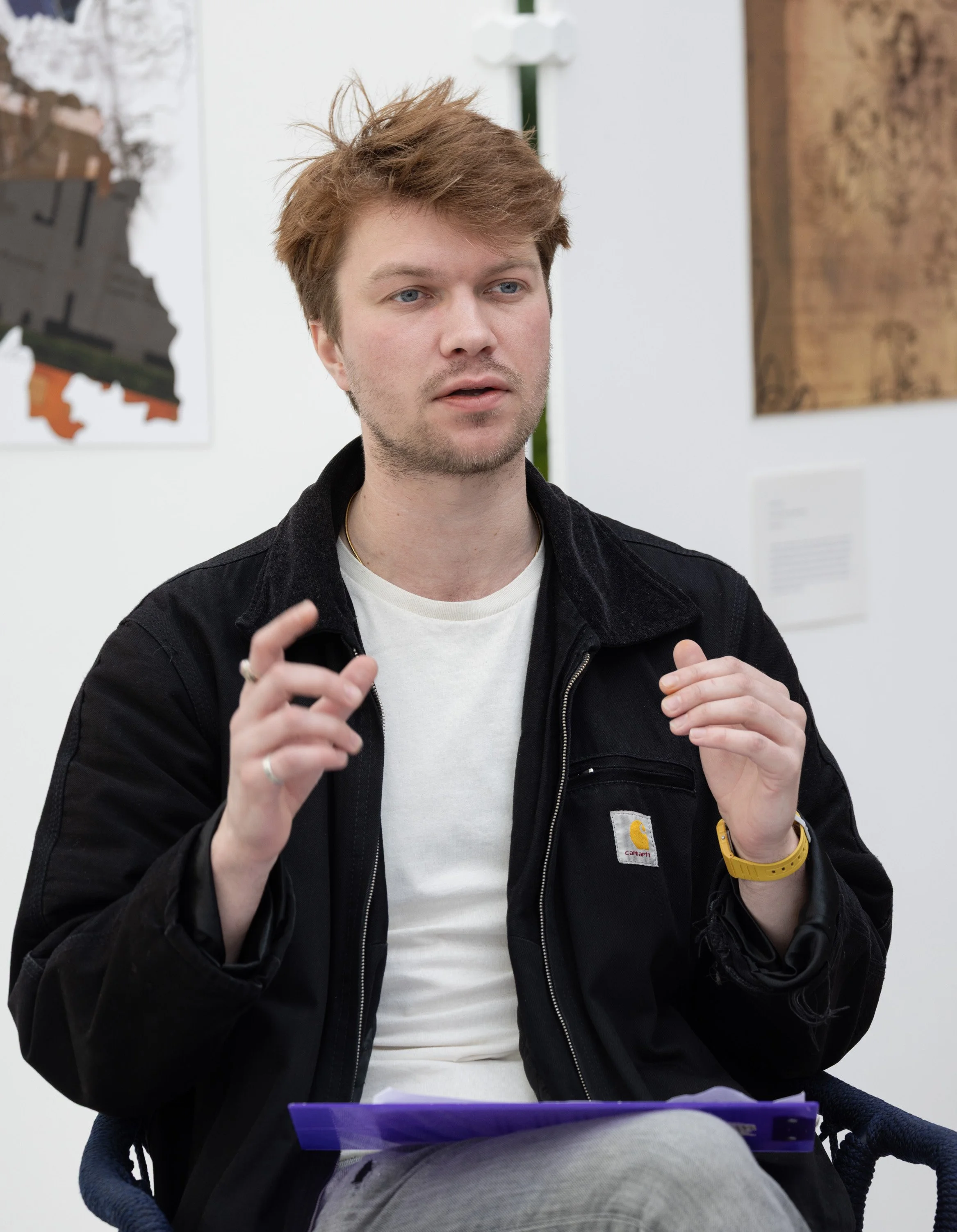 A young man with light brown hair, wearing a black Carhartt jacket, white t-shirt, and yellow wristwatch, sitting in a gallery or exhibit space, speaking or explaining something.