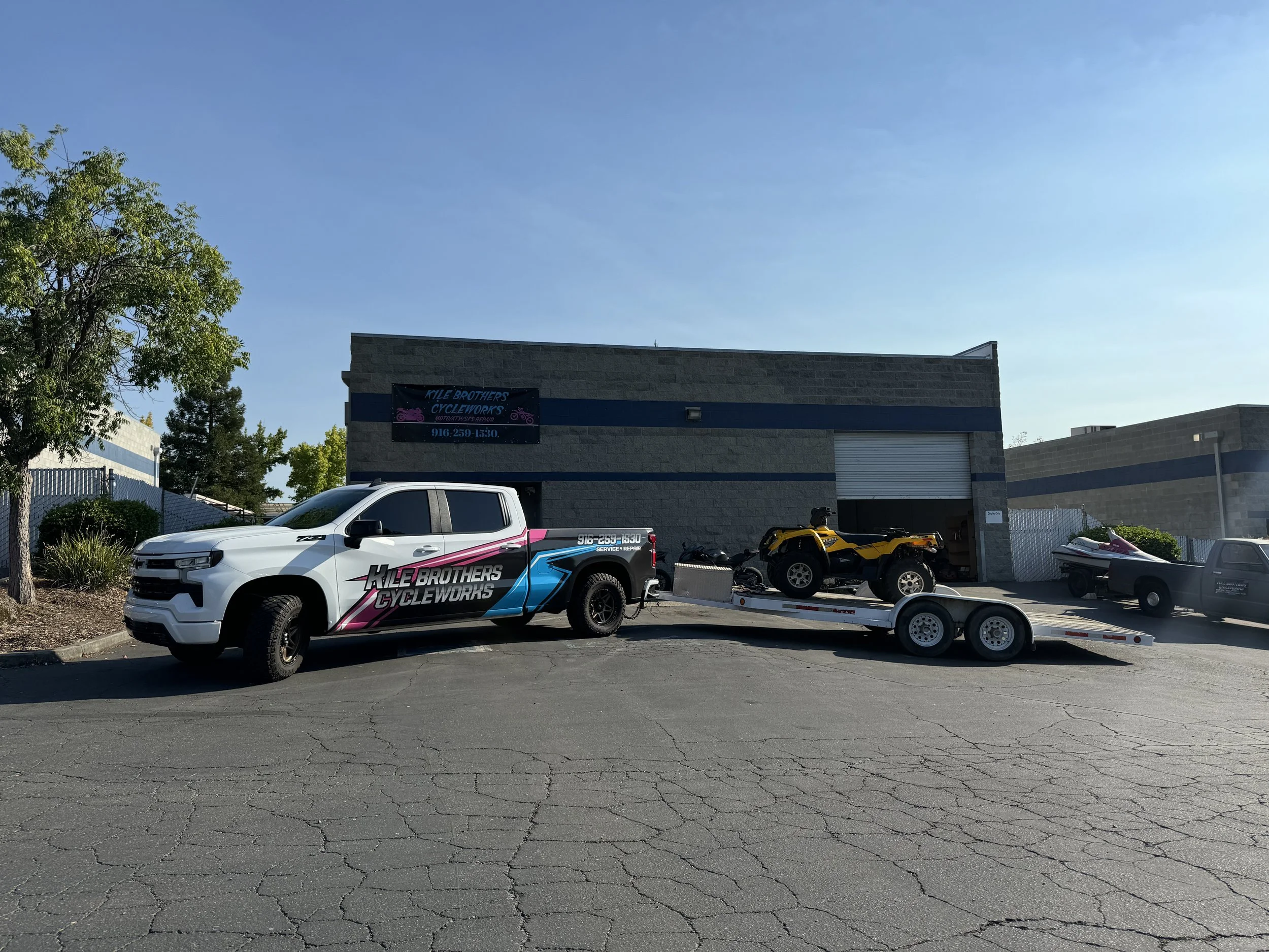 A white towing truck with 'KLE BROTHERS CYCLEWORKS' branding on the side, pulling a trailer carrying a yellow quad ATV, parked outside a commercial building with a sign for cycle works.