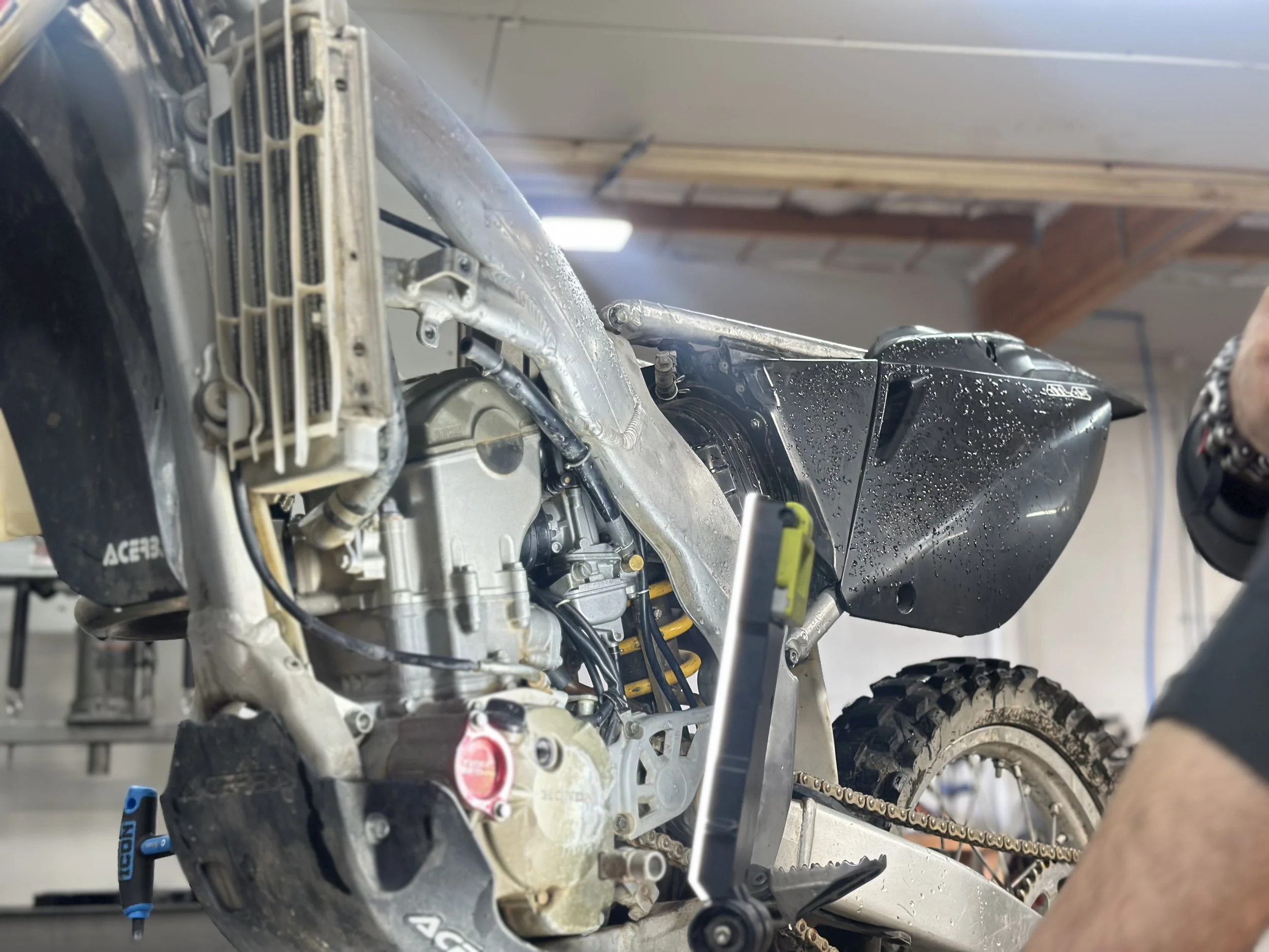 Close-up of a dirt bike's engine and front wheel, with water droplets on the black plastic part, in a workshop setting.
