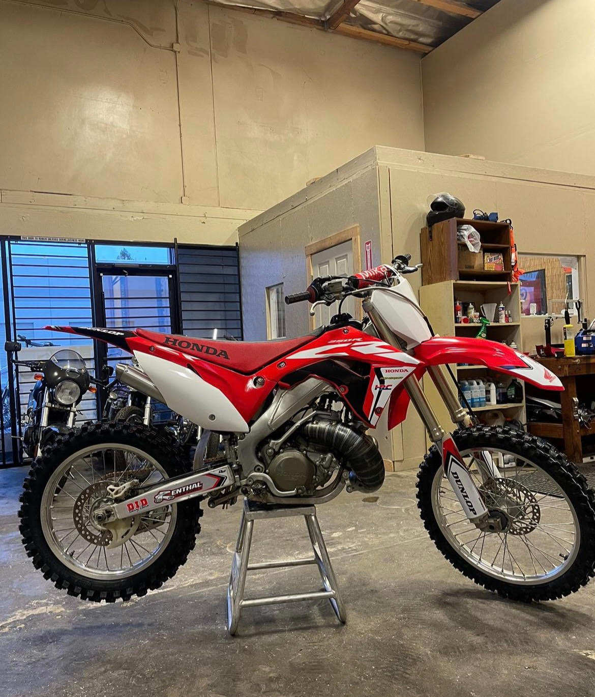 A red and white Honda dirt bike with knobby tires, parked on a small stand inside a garage or workshop with shelves, tools, and equipment in the background.