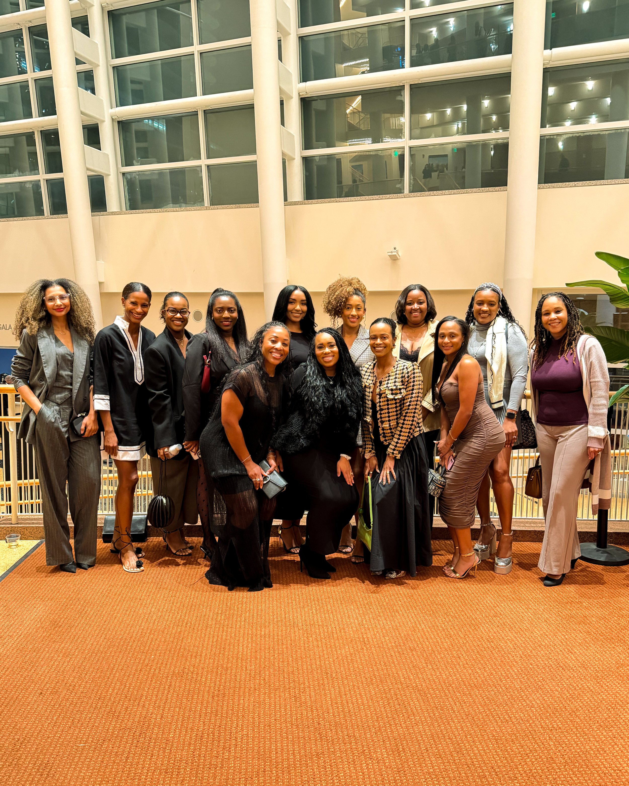 Group of 14 women posing for a photo at an indoor venue with large glass windows and warm lighting.