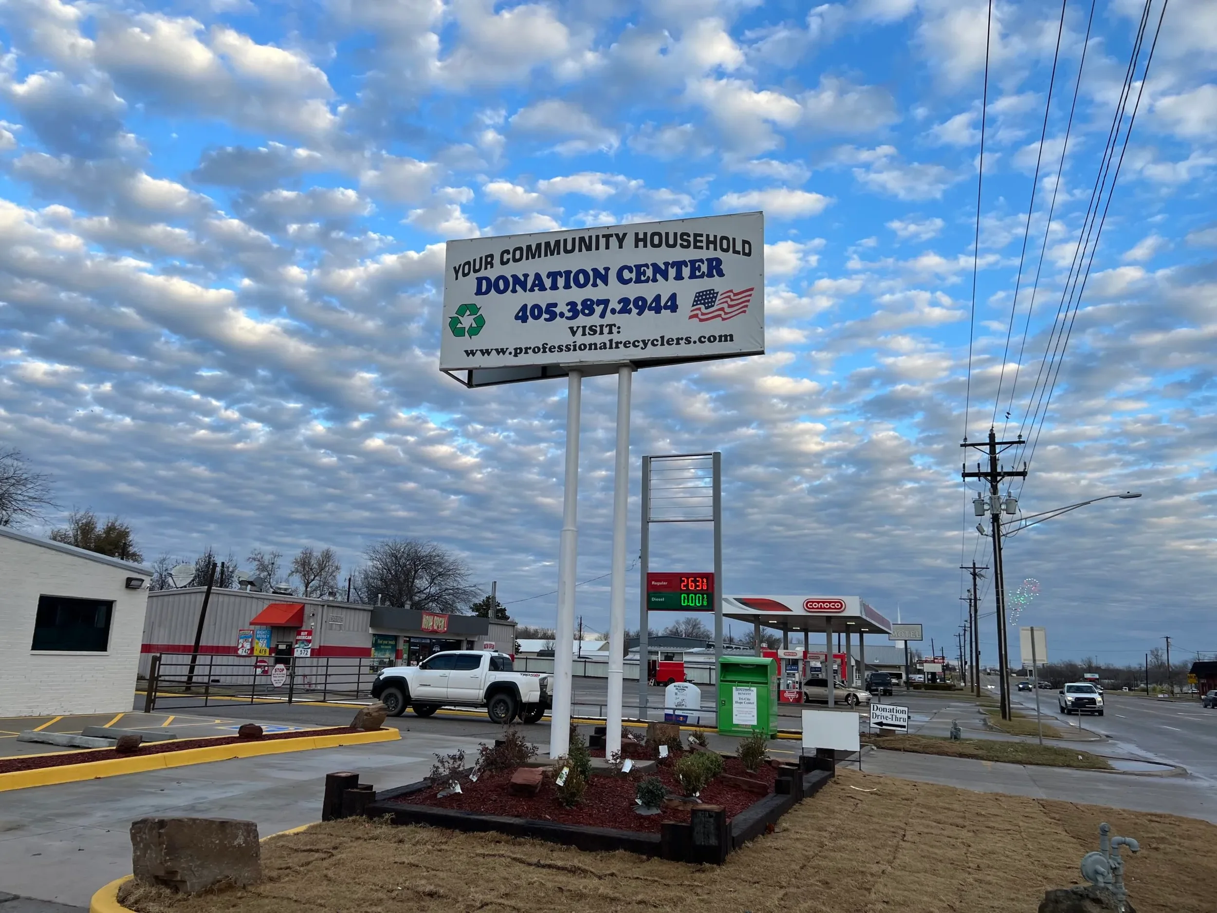 Billboard advertising community household donation center with contact number and website, in front of gas station and convenience store under a sky filled with clouds.