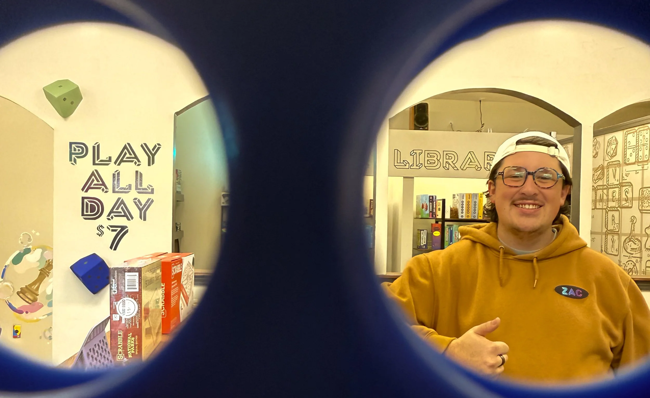 A young man with glasses and a white cap backwards, smiling and wearing a yellow hoodie, standing inside Very Good Game Store in front of the words 'GAME LIBRARY' on the wall in the background.