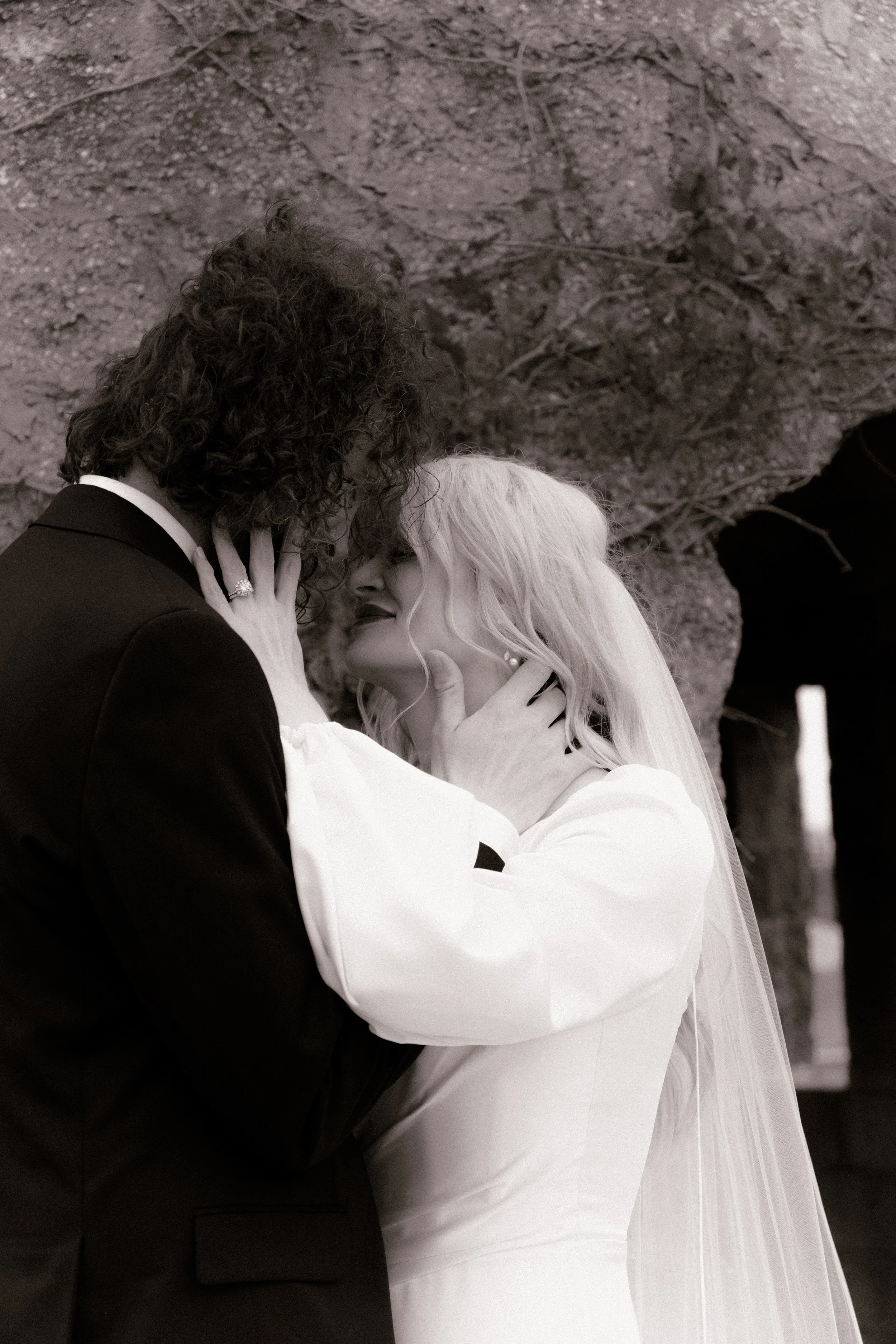 A bride and groom share a close, intimate moment during their wedding, with the bride wearing a white dress and veil, and the groom in a dark suit, touching each other's faces under a natural rock formation.