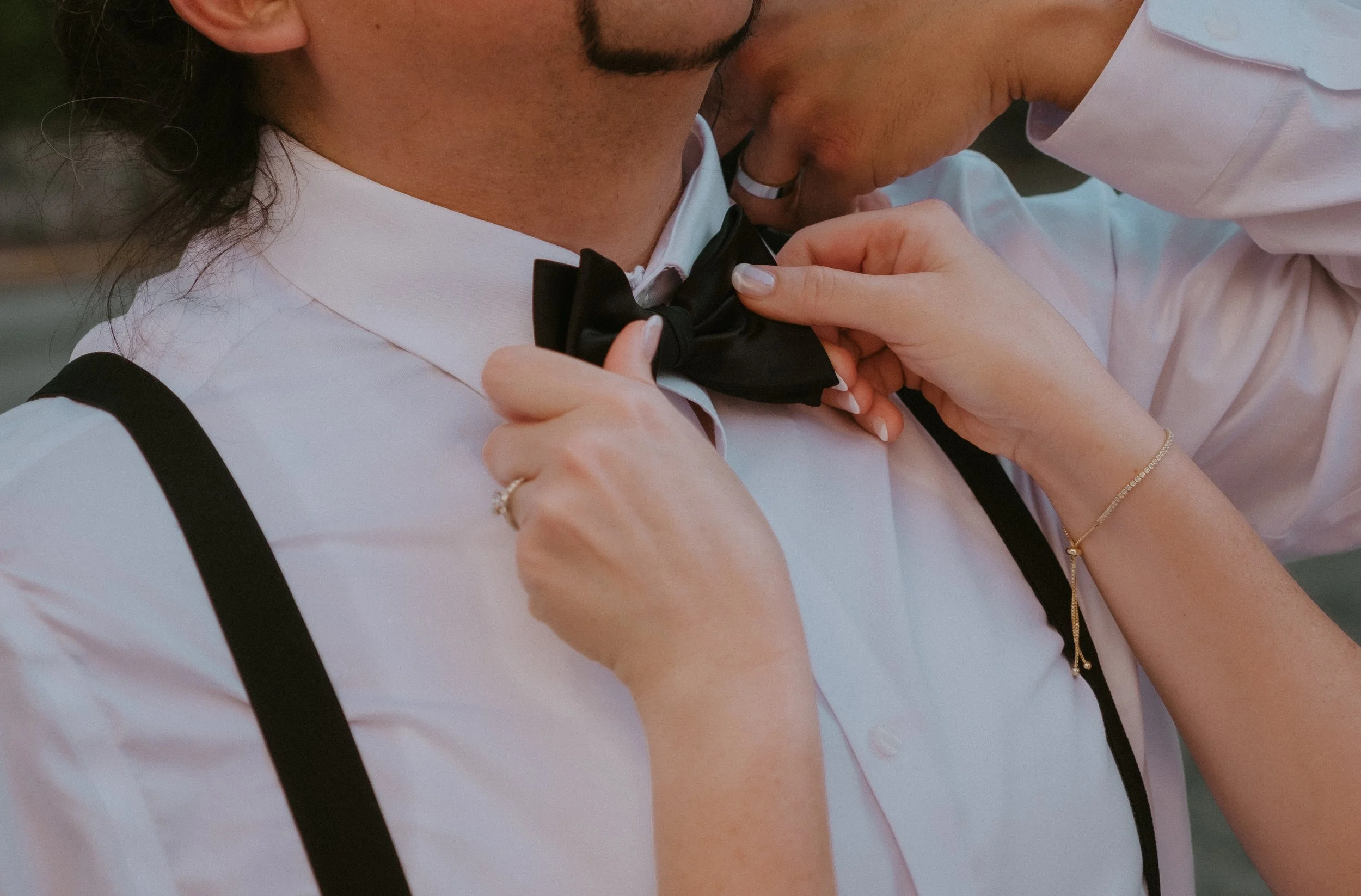 A woman adjusting a black bow tie on a man wearing a white dress shirt and suspenders.