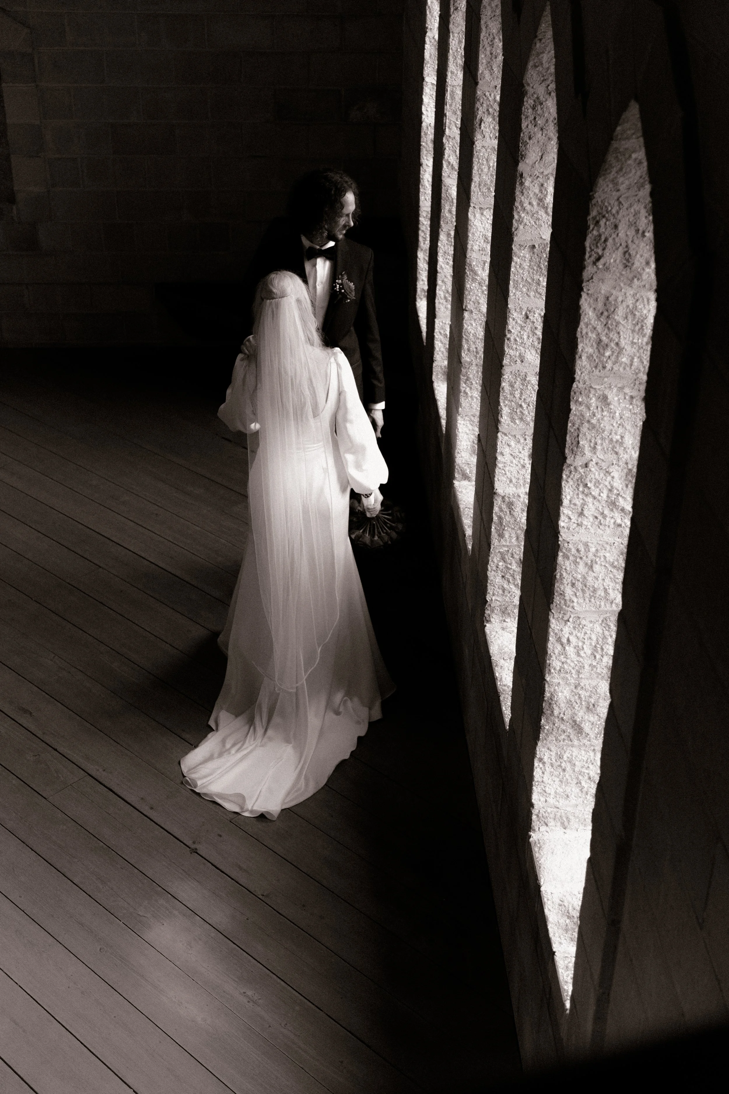 A black-and-white photo of a bride and groom standing together in an indoor setting, with the bride facing away and the groom looking at her, near a textured stone wall.