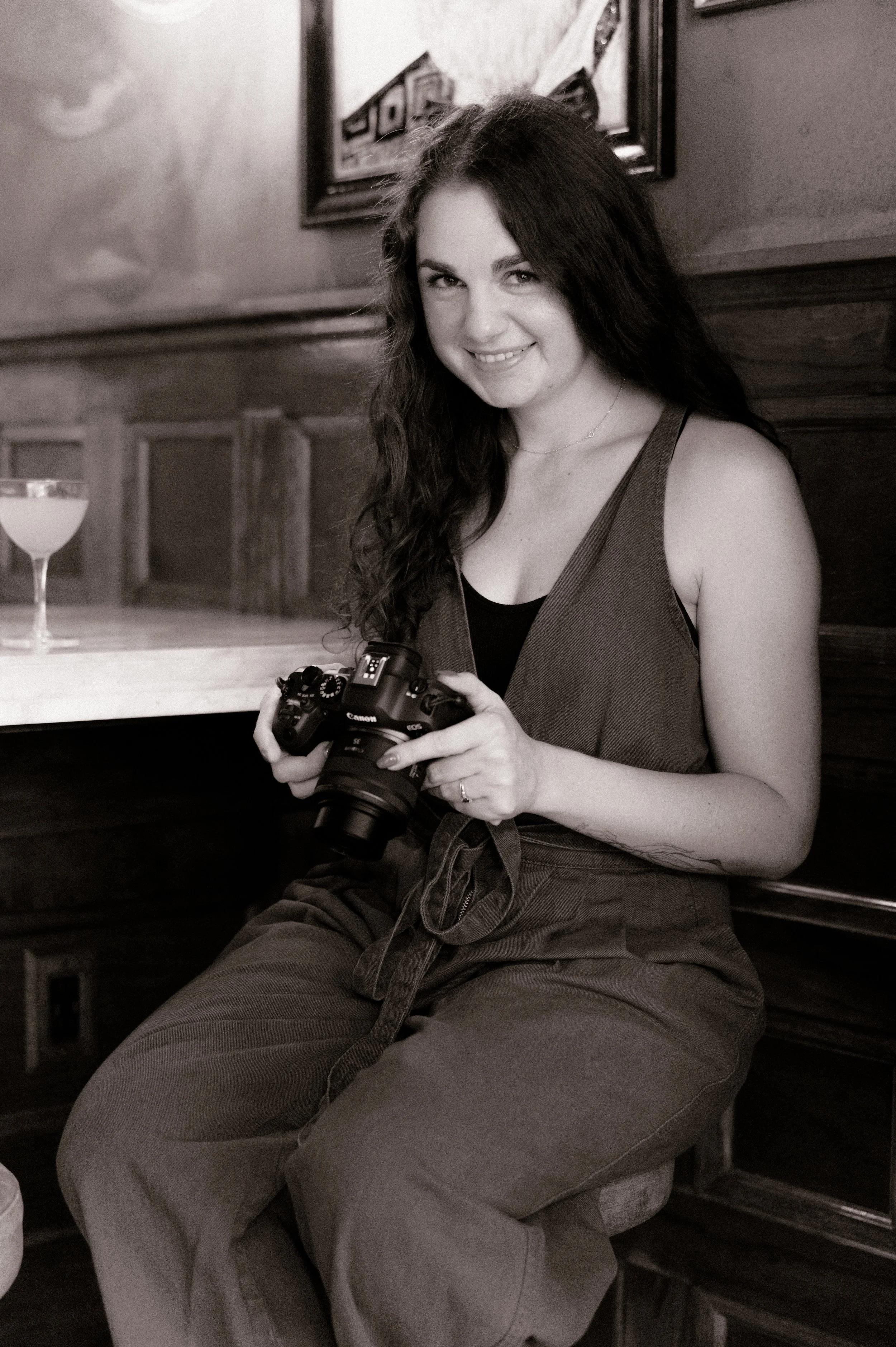 A woman with long, curly hair, smiling, holding a camera, sitting in a vintage-style restaurant with wooden decor.
