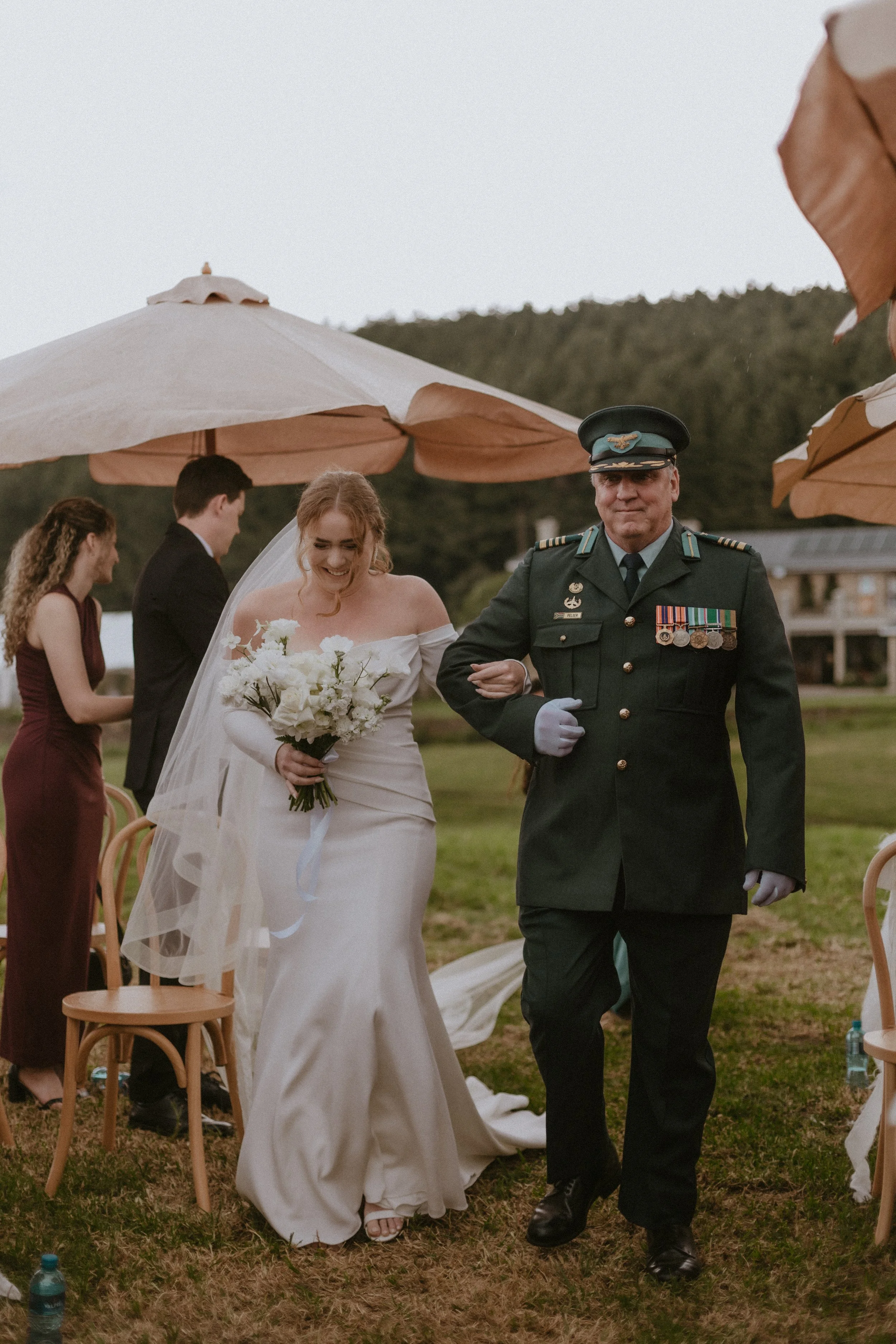 A bride in a white wedding dress holding a bouquet walks arm-in-arm with a uniformed military officer during an outdoor wedding ceremony.