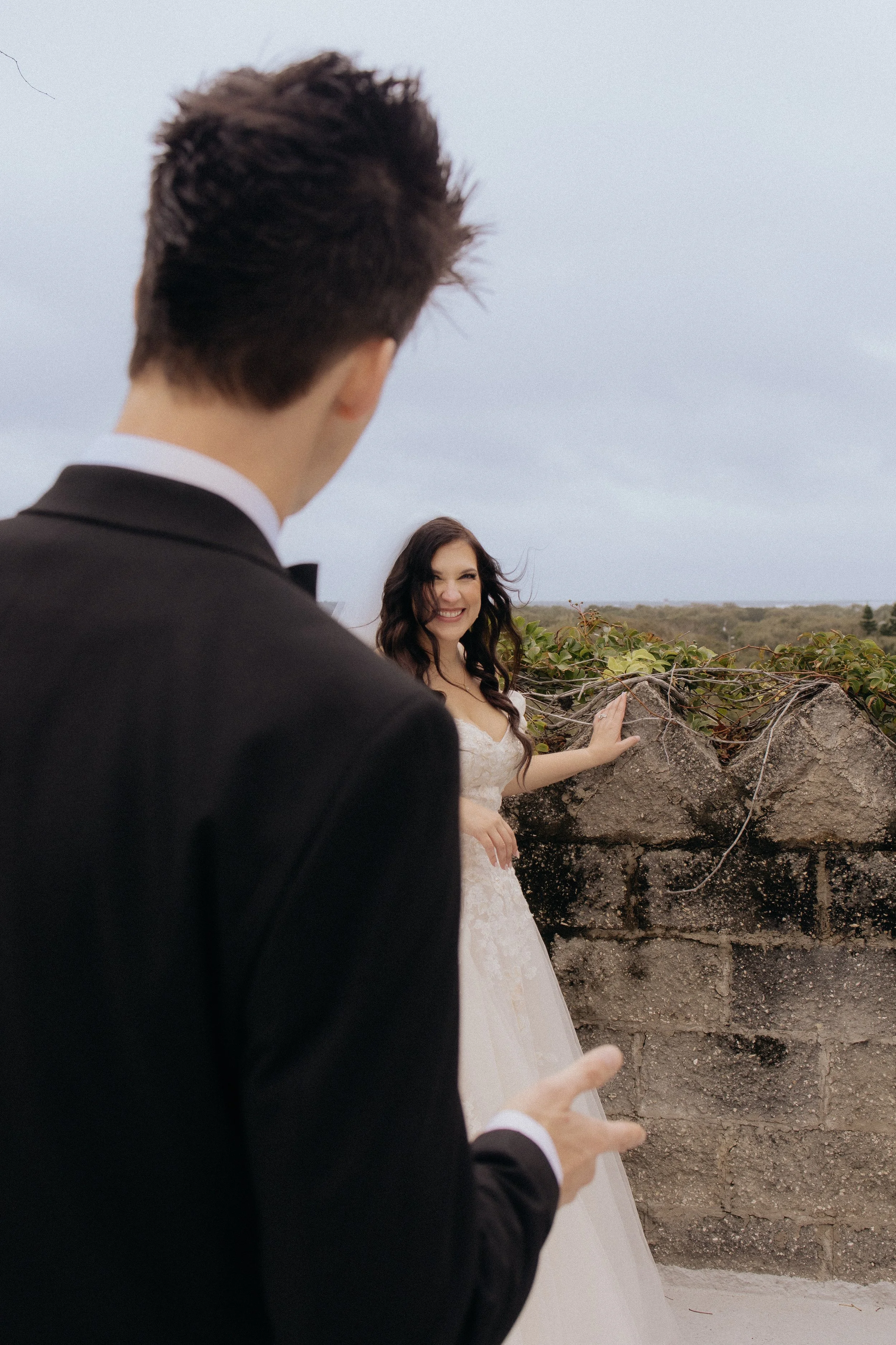 A man in a black tuxedo looks at a woman in a white wedding dress smiling on a rooftop with a stone wall and open landscape in the background.
