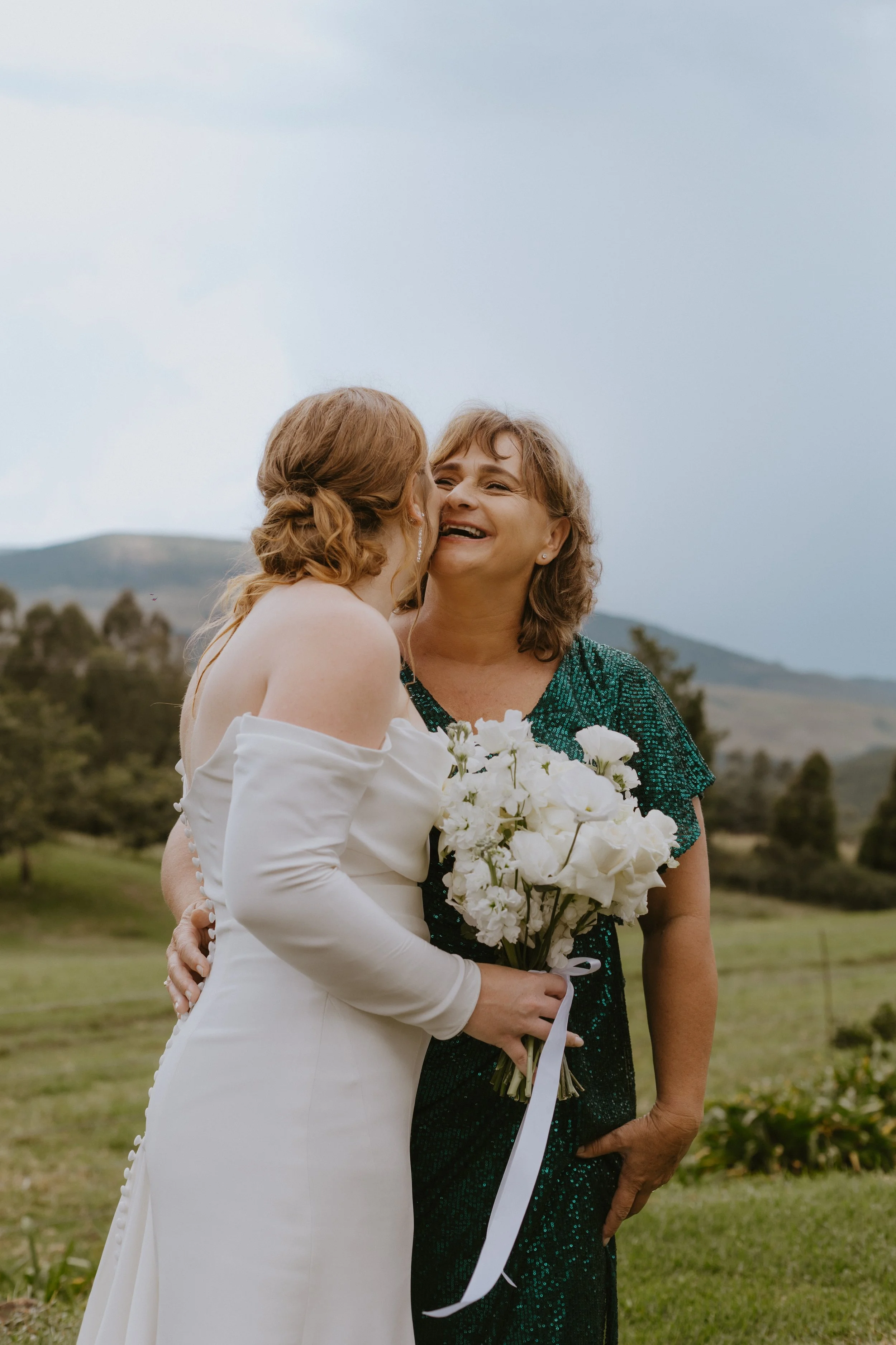 A bride and mom embracing outdoors, with the woman holding a bouquet of white flowers, smiling.