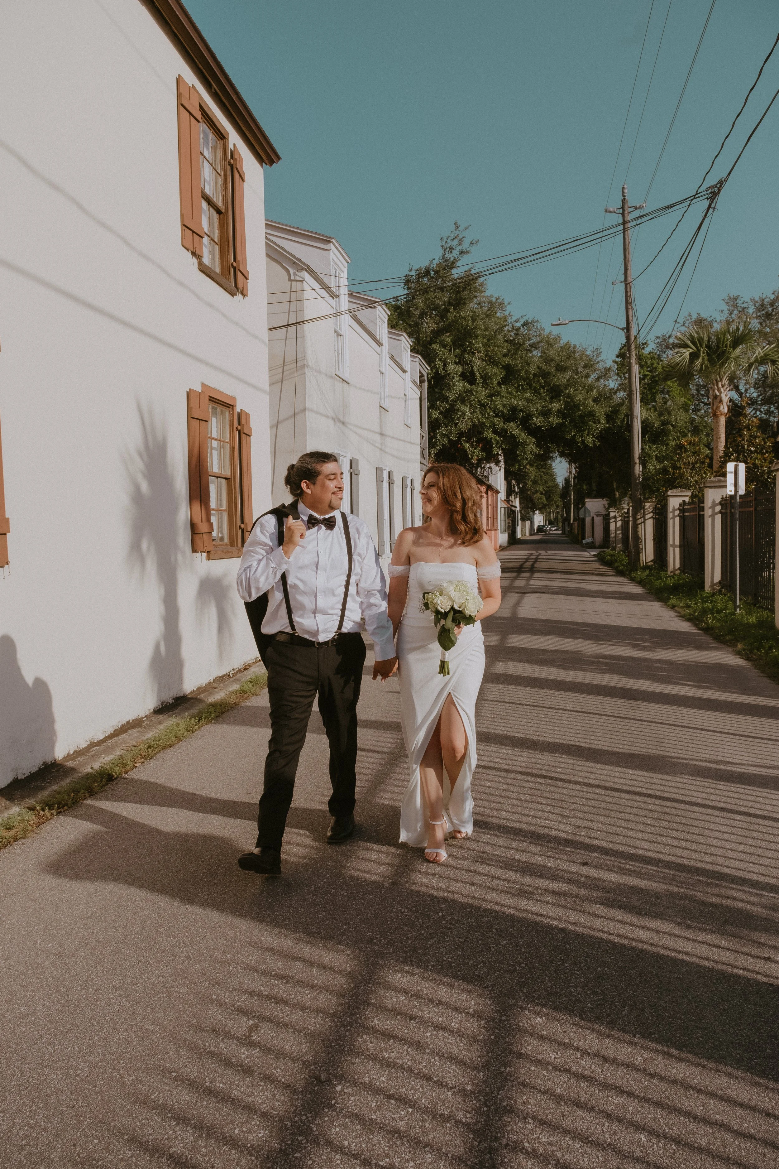 A couple in wedding attire holding hands and walking down a sunny street, with the bride carrying a bouquet and smiling at each other.