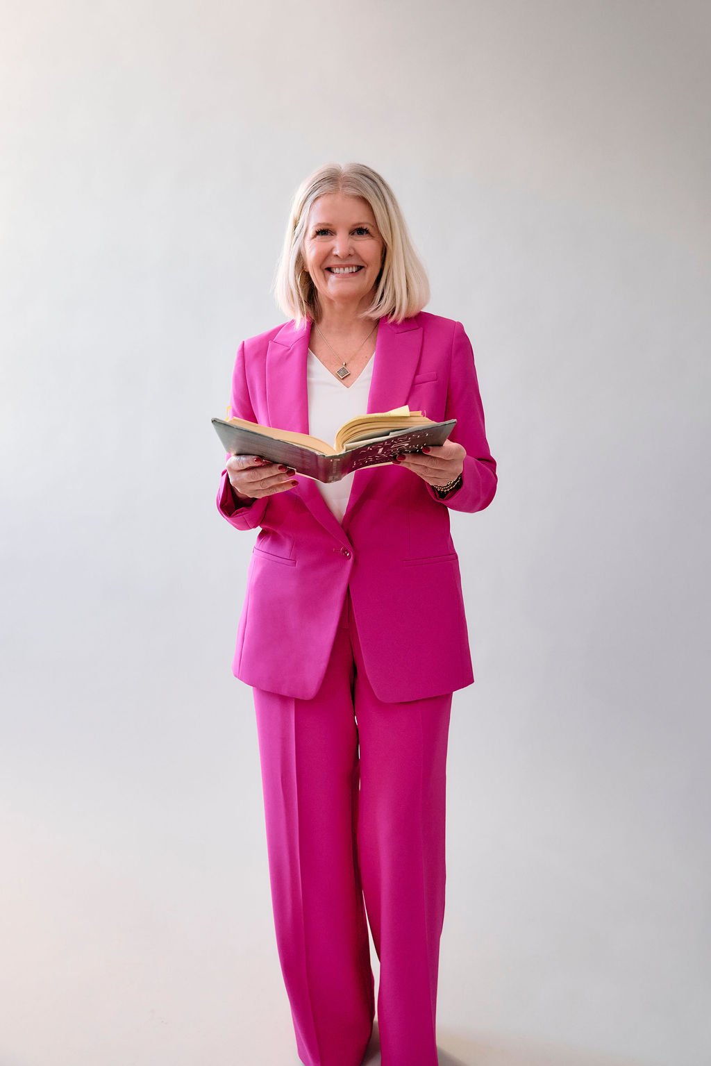 A smiling woman with blonde hair wearing a bright pink suit and white blouse, holding an open book, standing against a plain light-colored background.