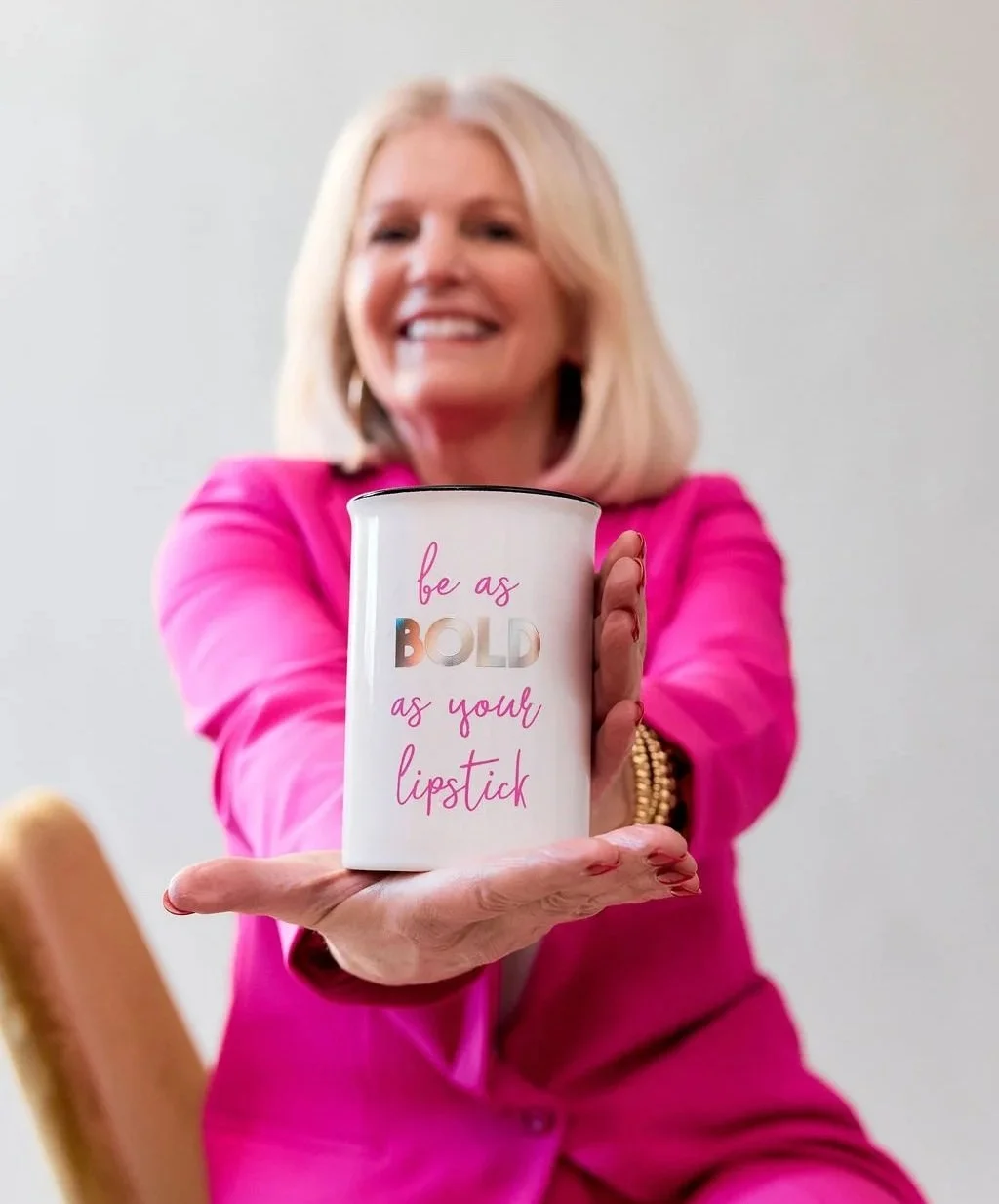A woman in a pink blazer holding a white mug with the phrase "be as BOLD as you, lipstick." The woman is smiling and looks happy.