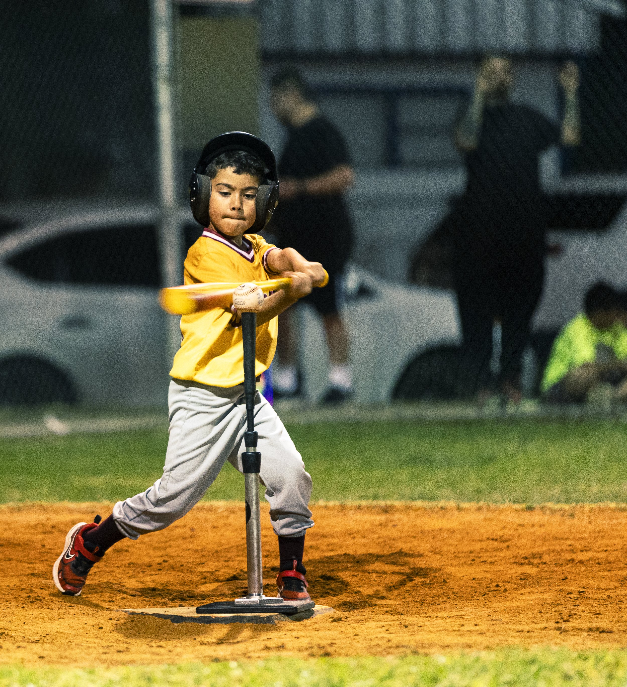 A young boy in a yellow sports jersey and gray pants practicing baseball hitting with a tee, wearing protective headphones, on a baseball field at night.