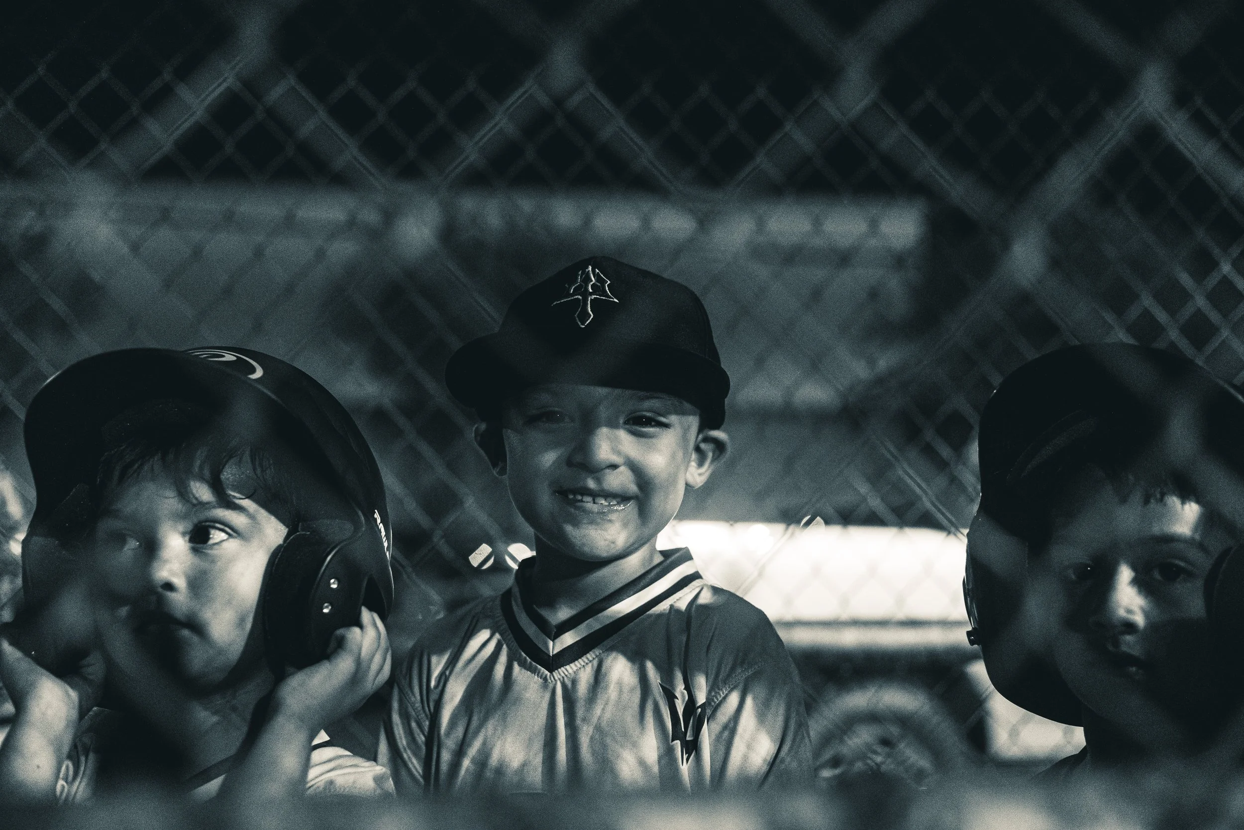 Three young boys at a baseball game, wearing helmets and smiling, seen through a chain-link fence.