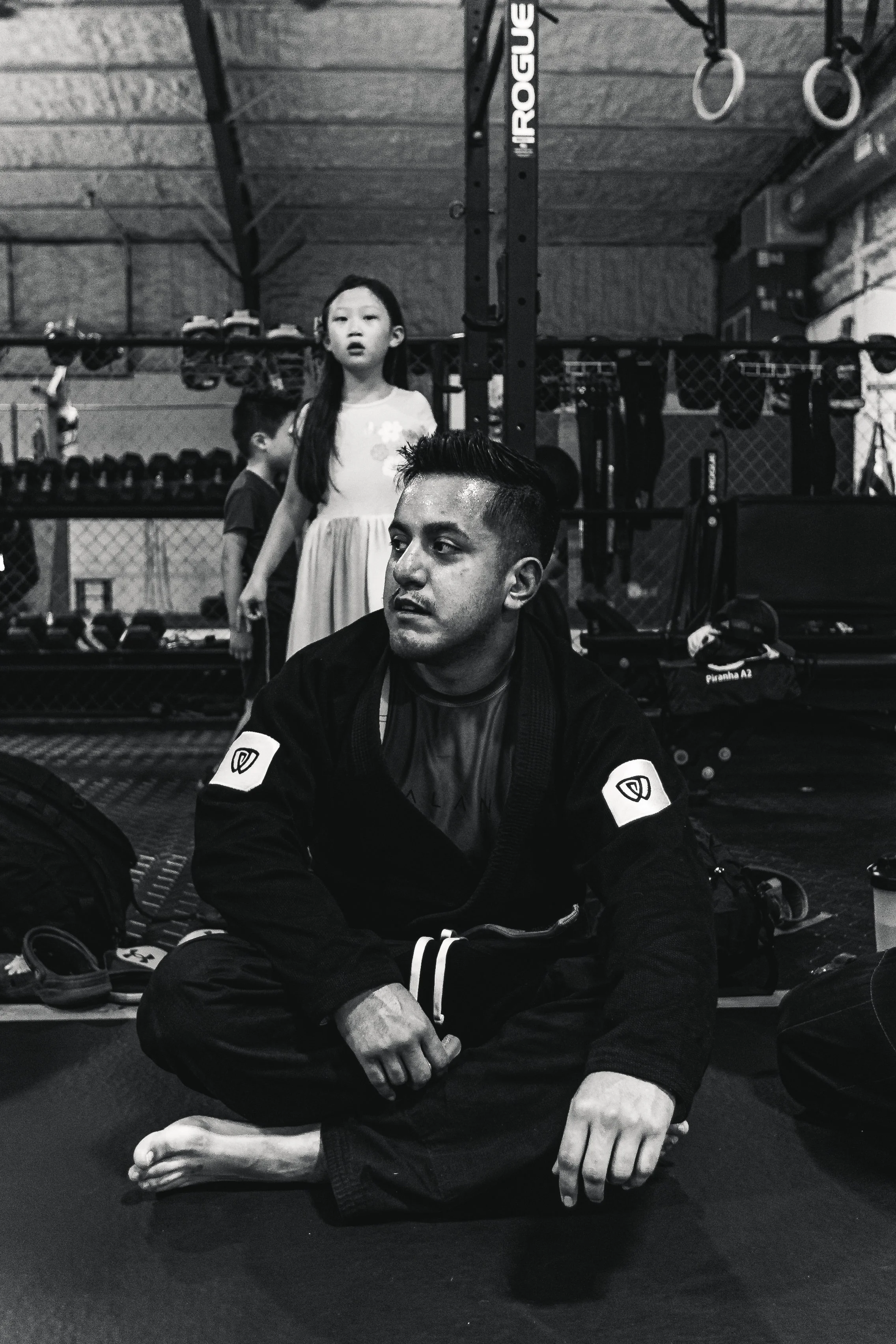 A man in a martial arts gi sitting cross-legged on a black mat in a gym, with children and gym equipment in the background.