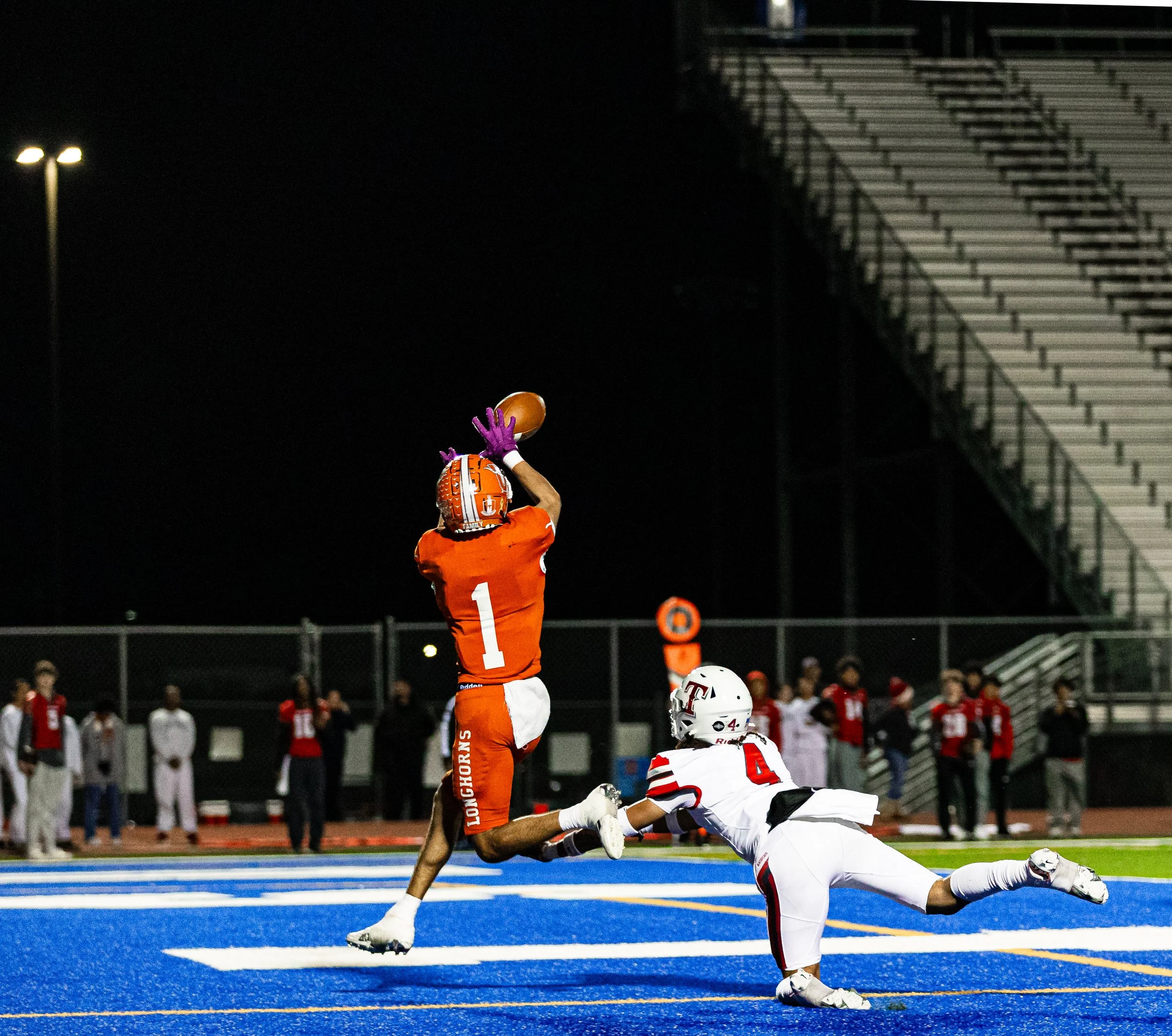 Football player in orange uniform catching a football in the end zone while an opposing player in a white uniform dives to block.