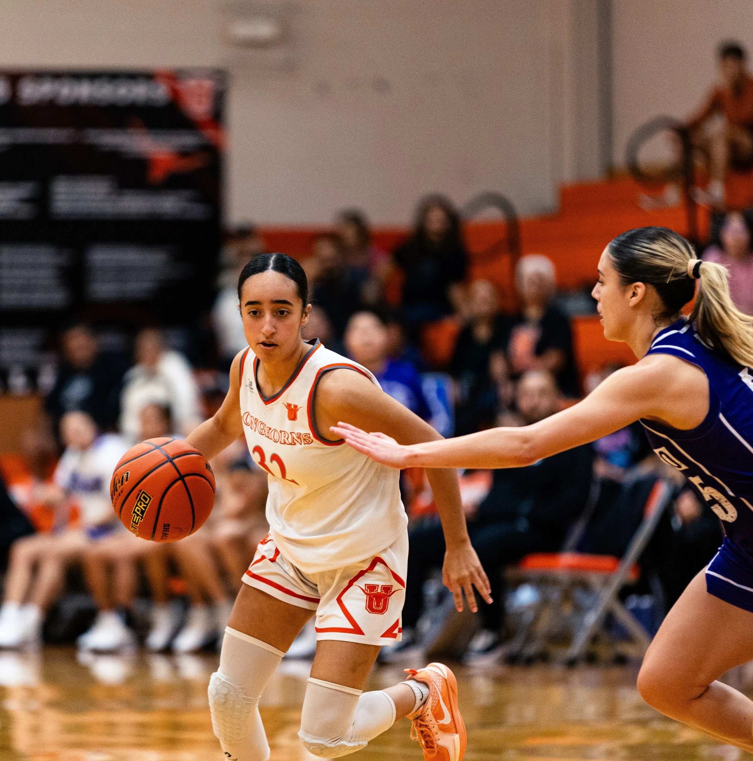 A female basketball player in a white and orange uniform dribbling an orange basketball while being guarded by a female opponent in a blue uniform during a game in a gymnasium filled with spectators.