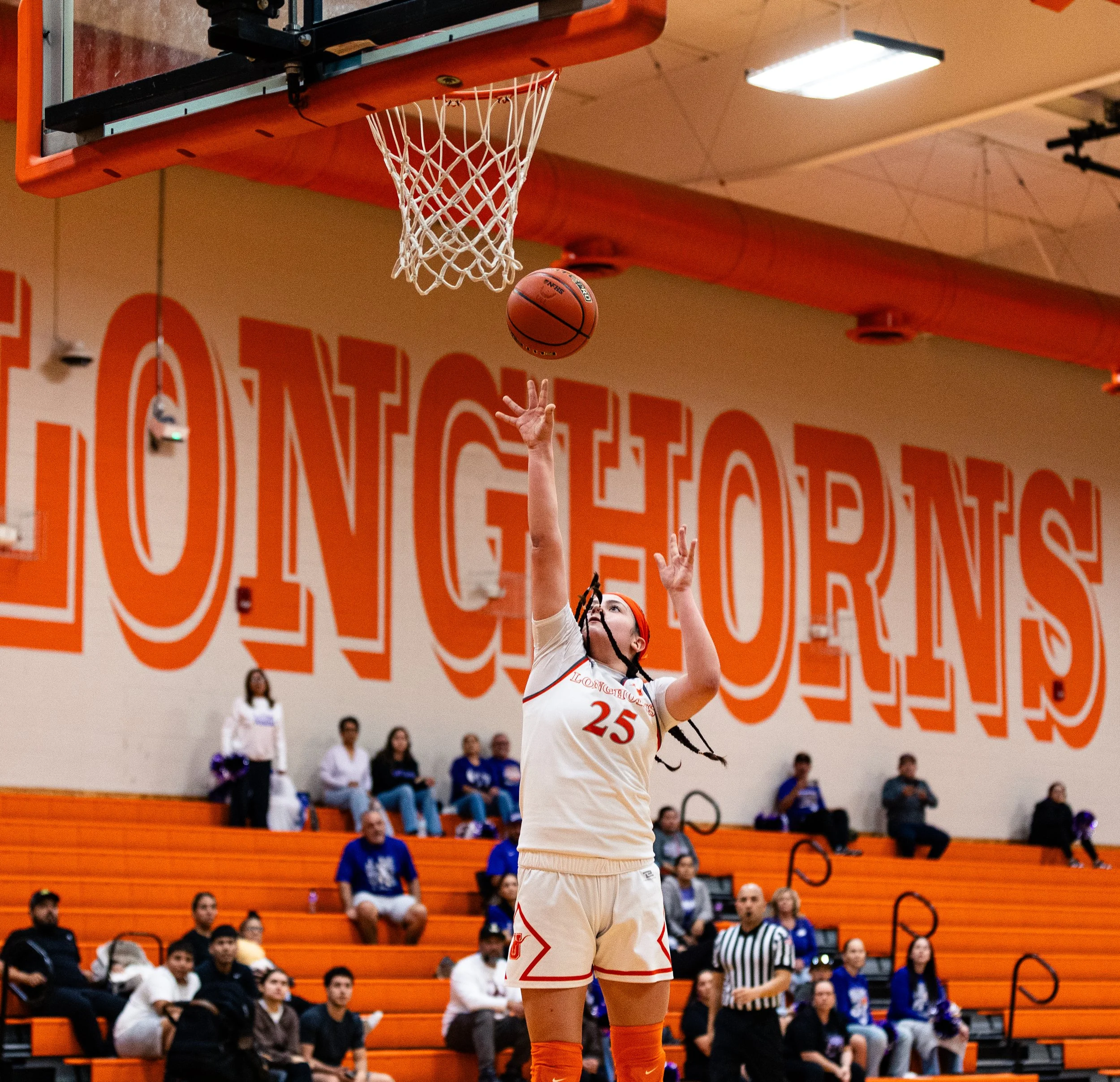 A female basketball player wearing a white jersey with the number 25, orange shorts, and an orange headband is shooting a basketball towards the hoop in a gymnasium. The gym has orange seating and orange wall decorations, with spectators watching the
