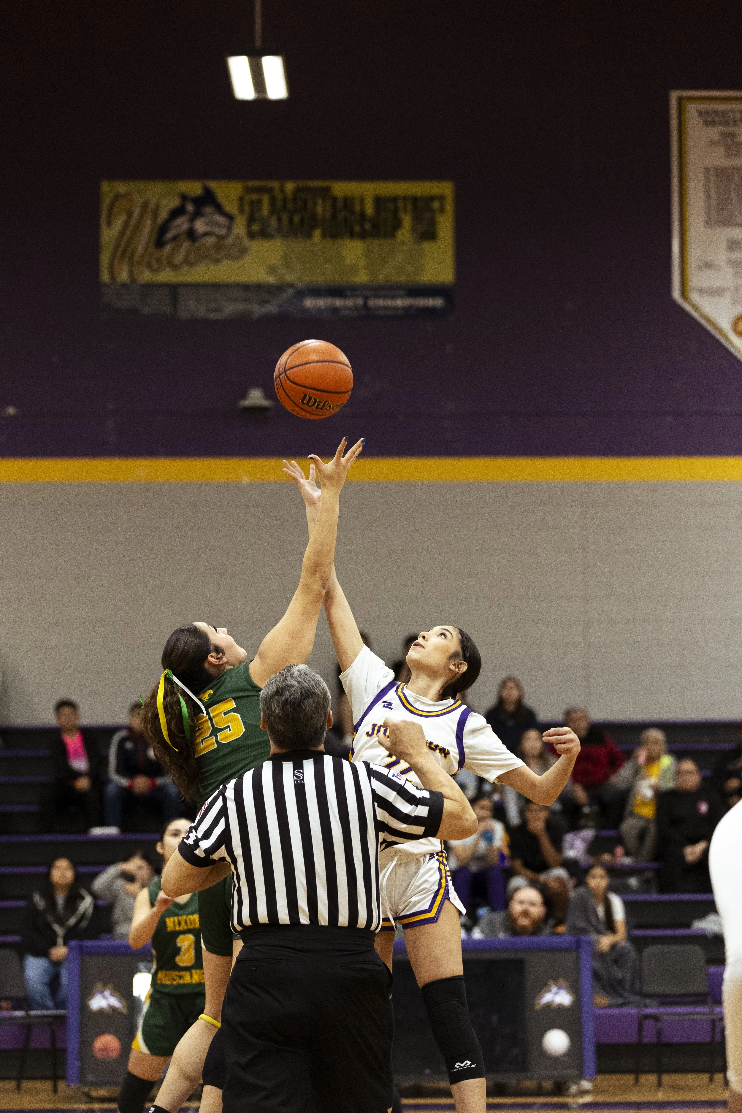 Two female basketball players are jumping for the ball during a game, with a referee watching. One player in green and yellow and the other in white and purple are contesting the ball at a tip-off. Spectators are seated in the background.