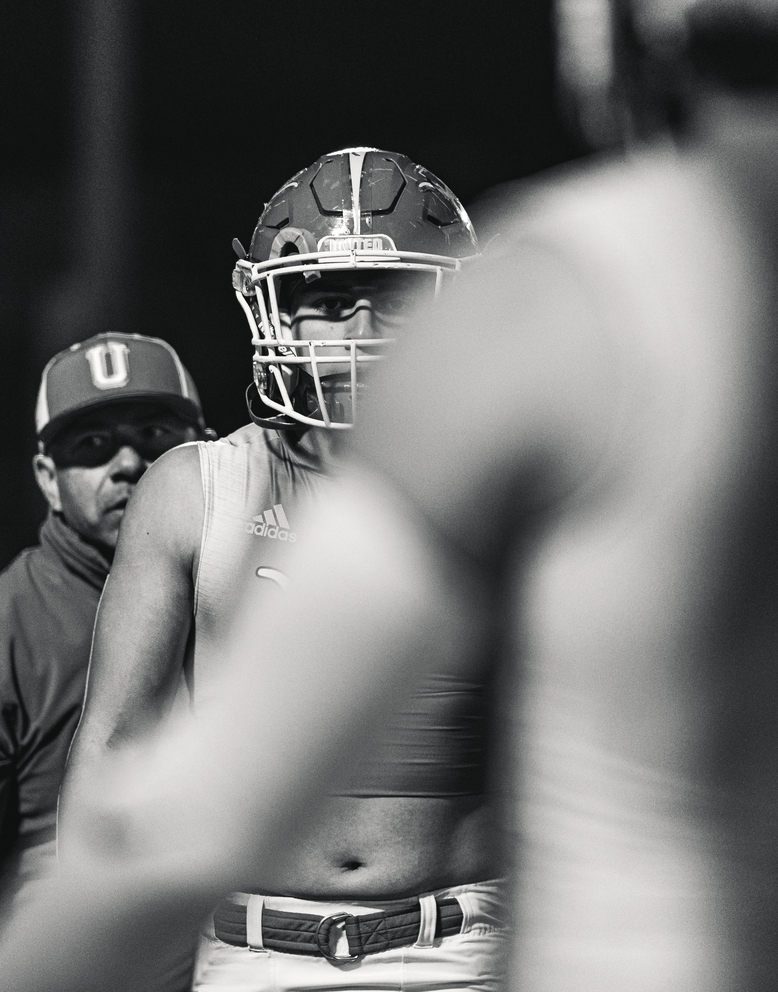 Black and white photo of a football player wearing a helmet, sleeveless shirt, and athletic gear, with a person in the background.