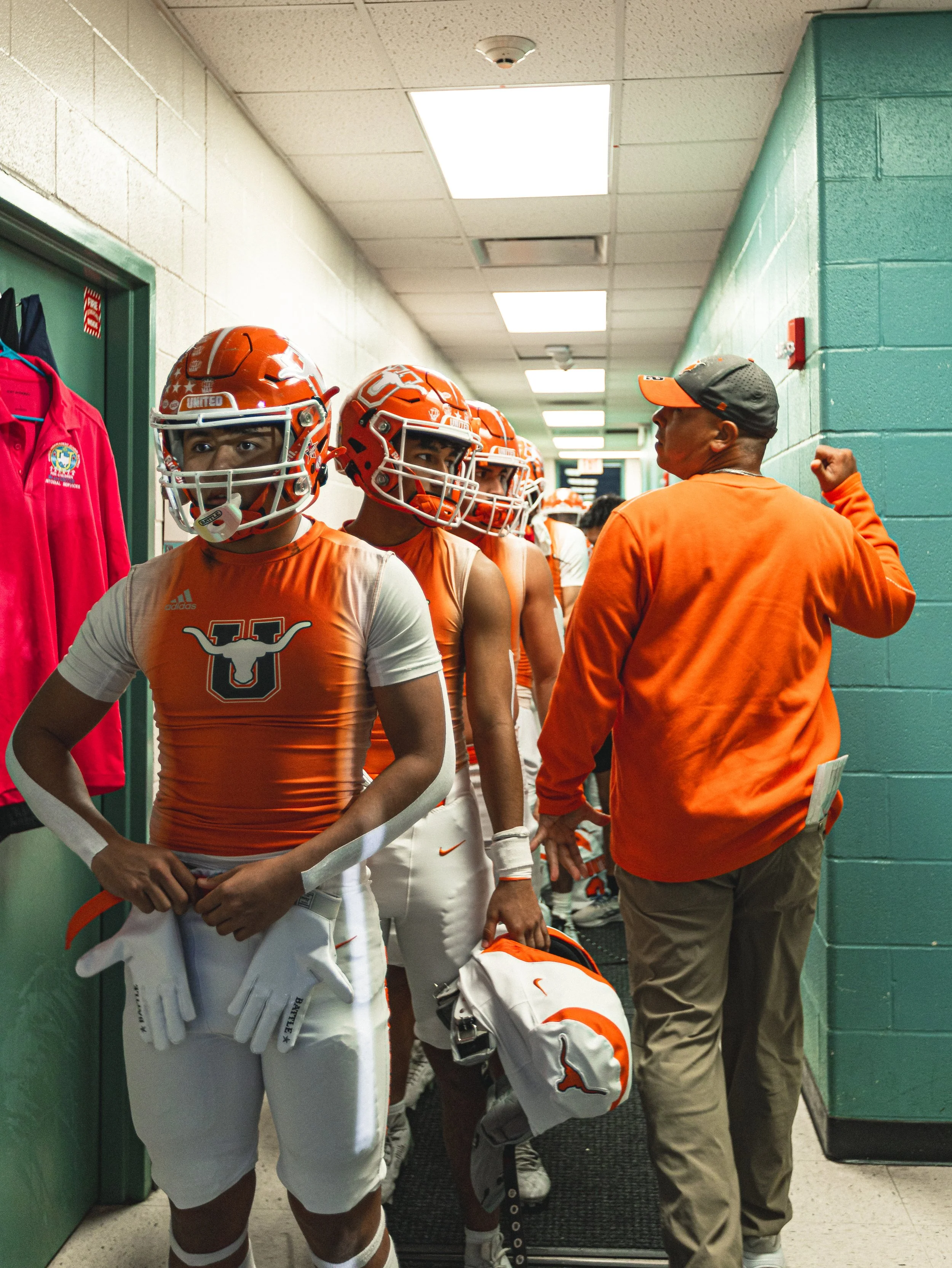 Football players in orange and white uniforms with helmets, historic locker room with teal walls, coach giving instructions.