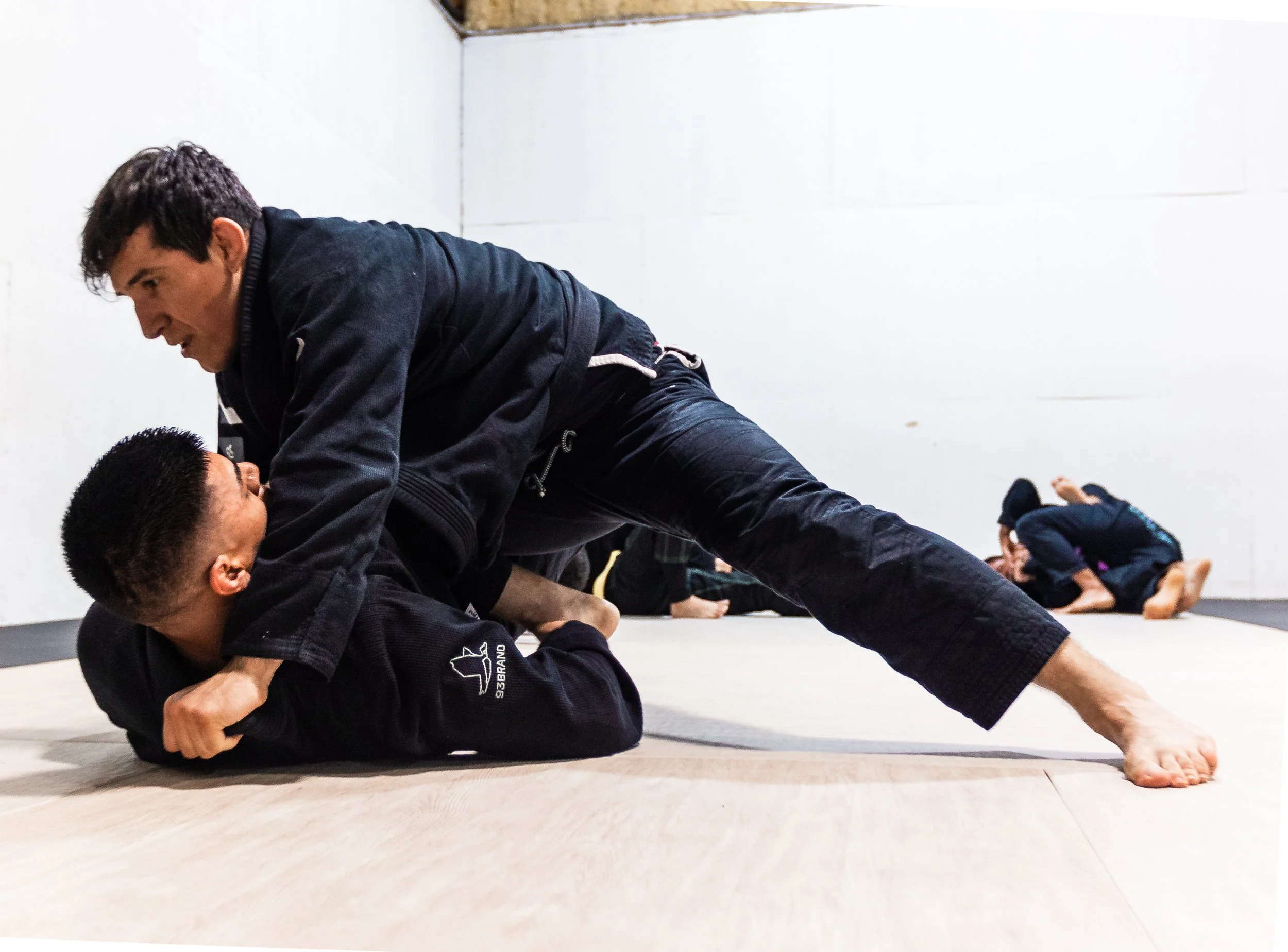 Two men practicing Brazilian Jiu-Jitsu on mats, with other practitioners in the background.